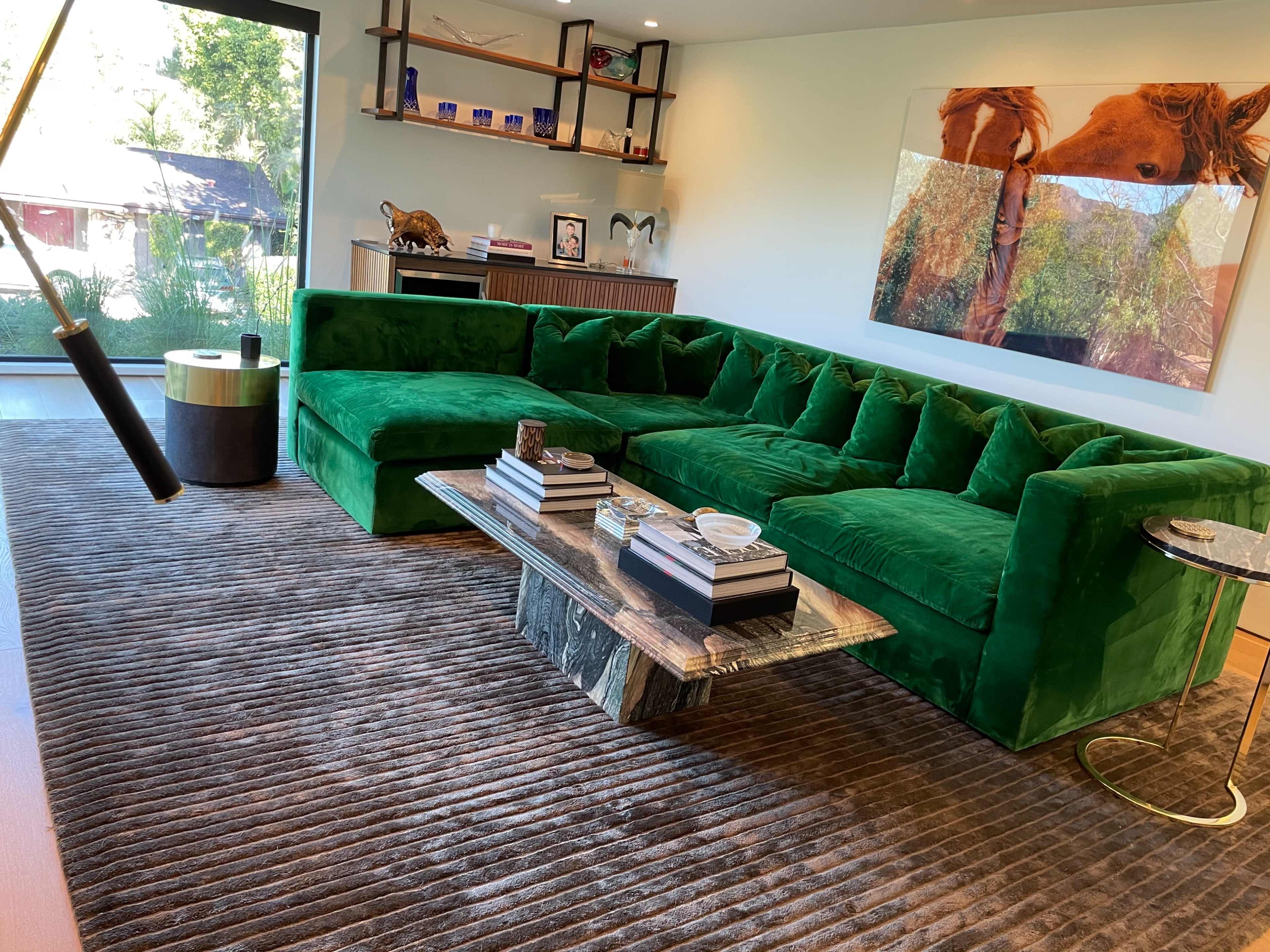 A modern living room featuring a large green velvet sectional sofa, a striped area rug, and a coffee table topped with books and a decorative bowl.