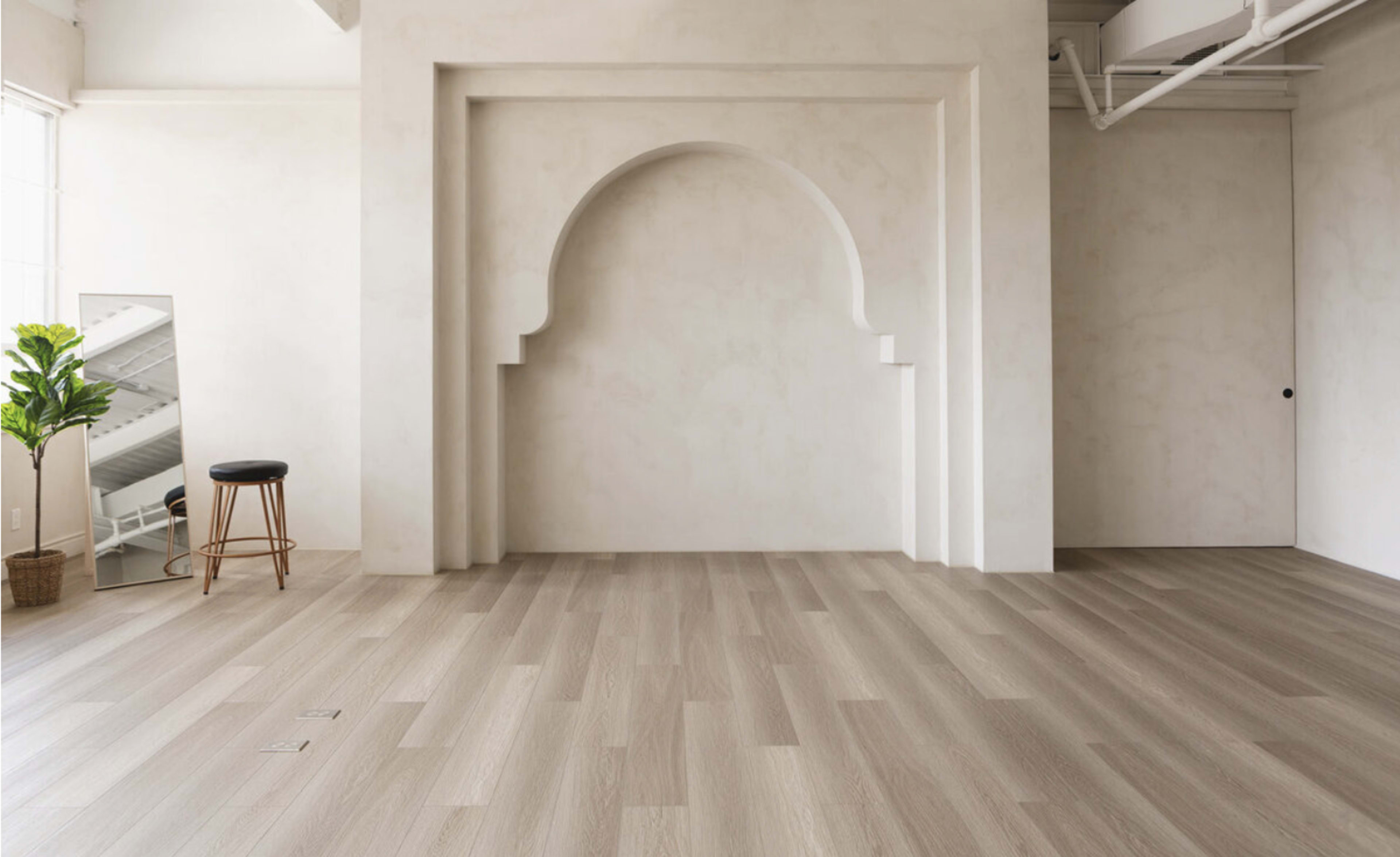 A spacious room with light-colored wood flooring, a large arched wall recess, a stool, a mirror, and a potted plant.