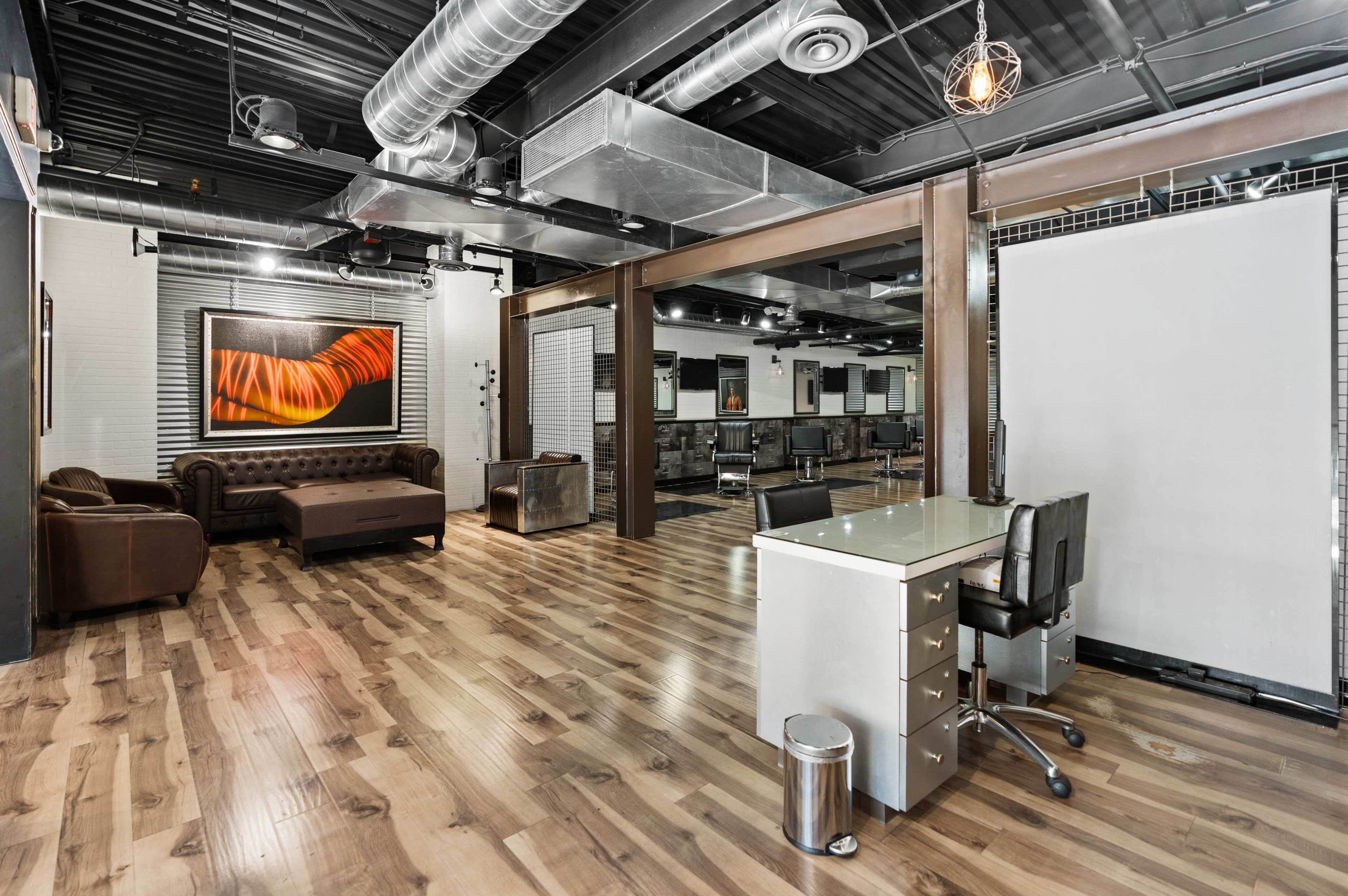 The image shows a modern, spacious interior with polished wooden floors, a brown leather couch, and an office desk with a rolling chair, framed by industrial-style decor.