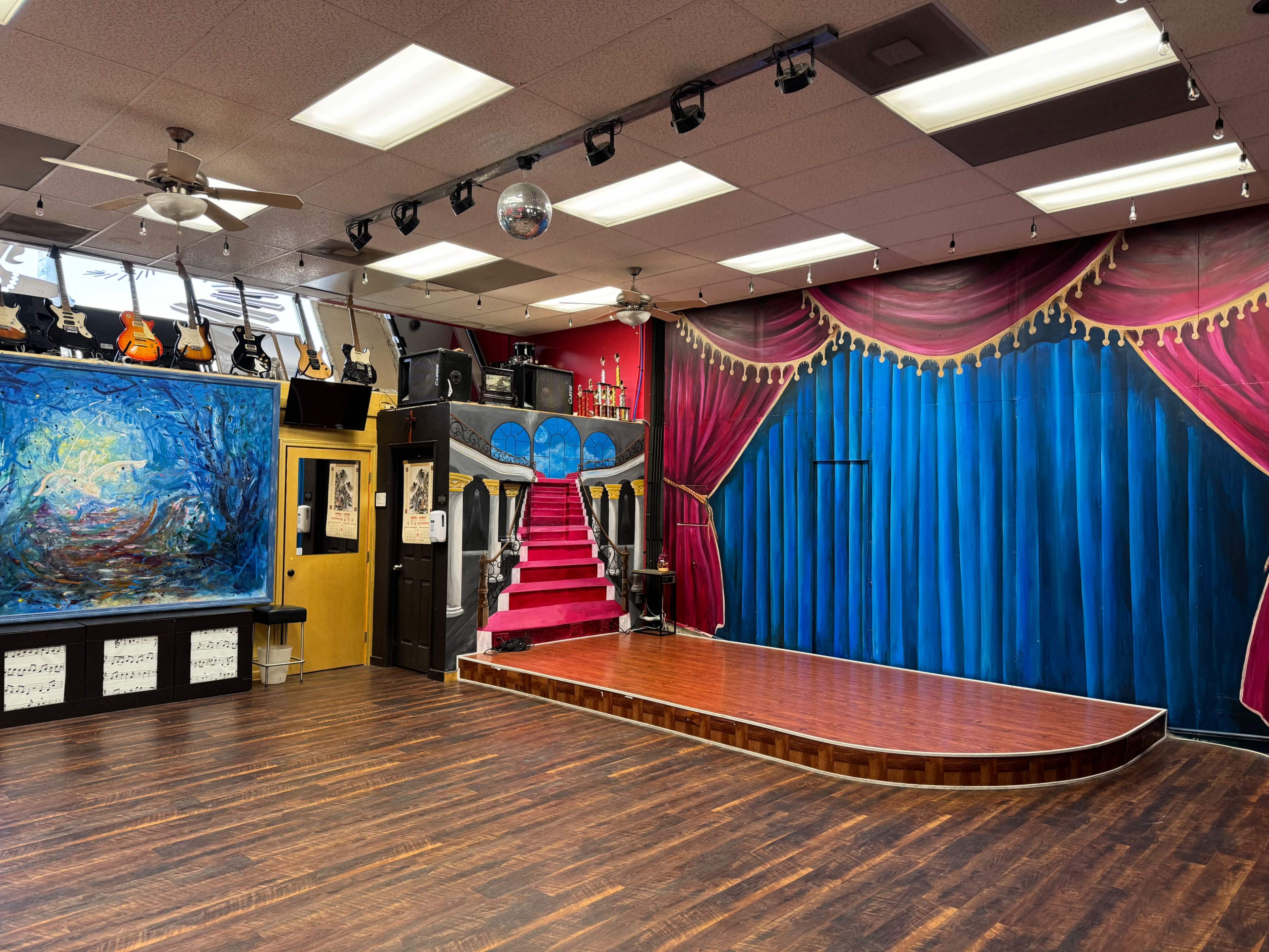 The image shows a performance space with a wooden stage, red-curtained backdrop, and a wall displaying musical instruments and artwork.