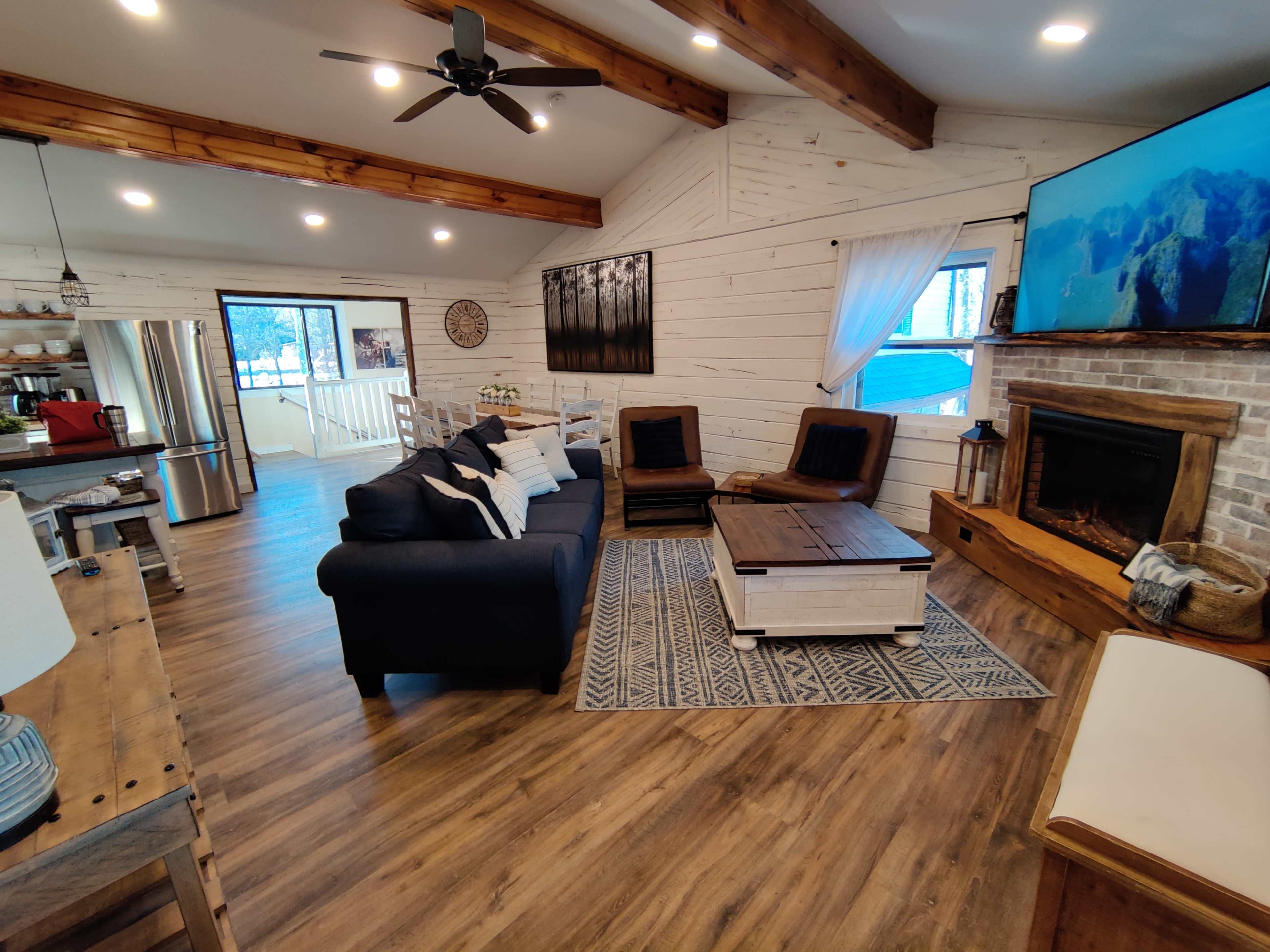 The image shows a cozy living room with a sectional couch, a coffee table, and a fireplace, featuring wooden beams and light-colored walls.