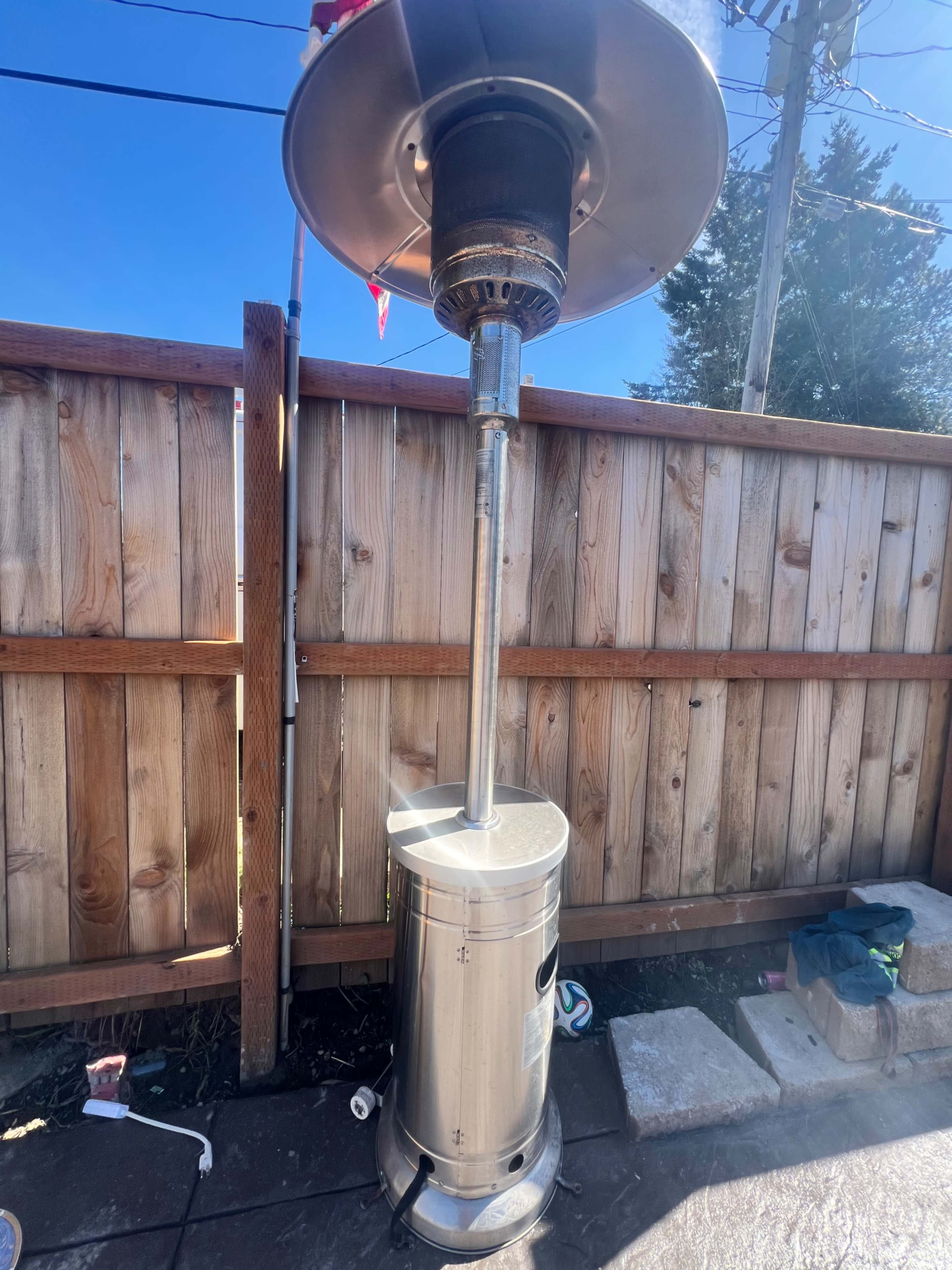 A stainless steel patio heater stands on a patio next to a wooden fence under a clear blue sky.