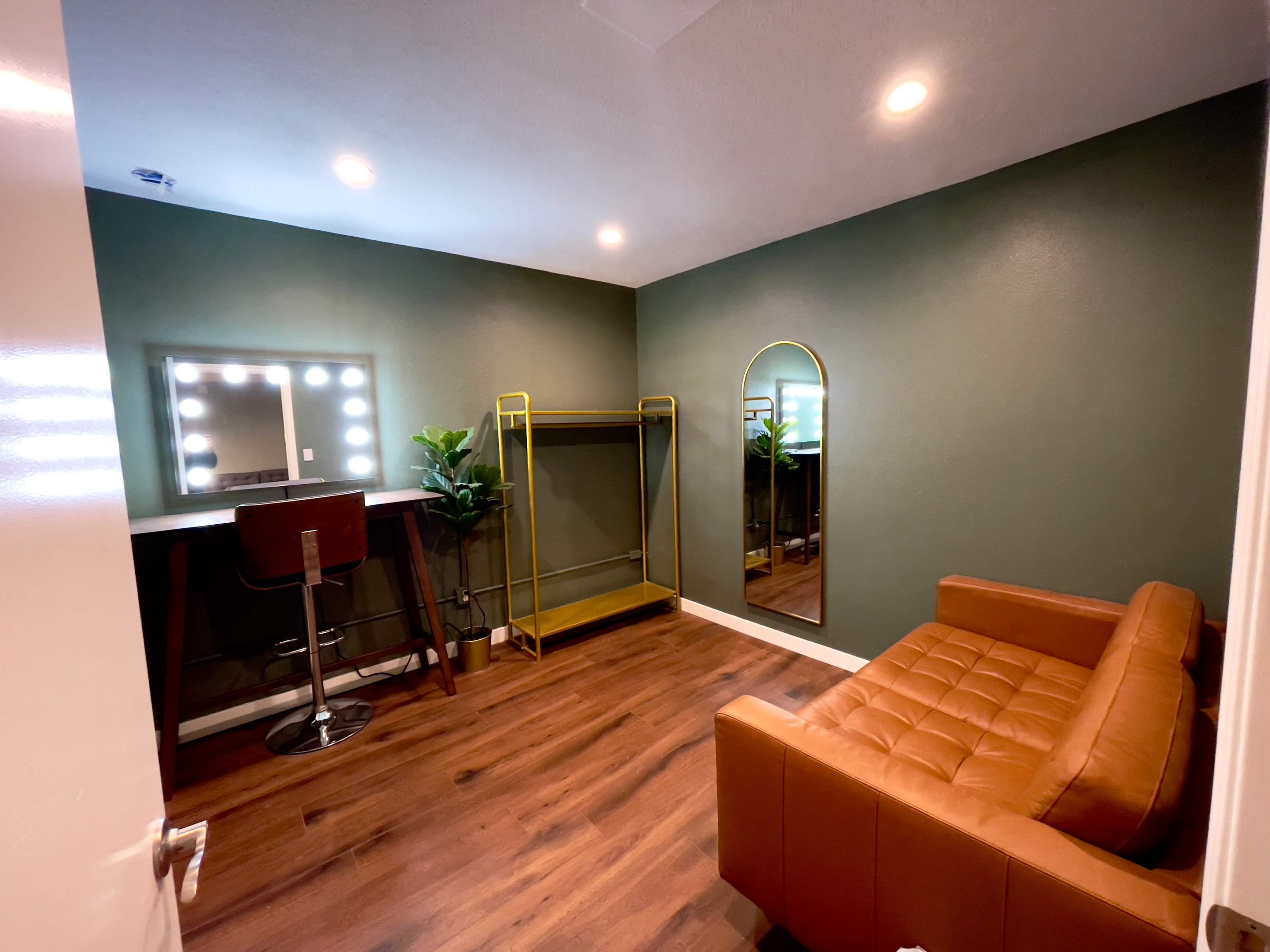 A small room with green walls, a mirror with lights, a tall gold clothing rack, a brown sofa, and a bar-style seating area.