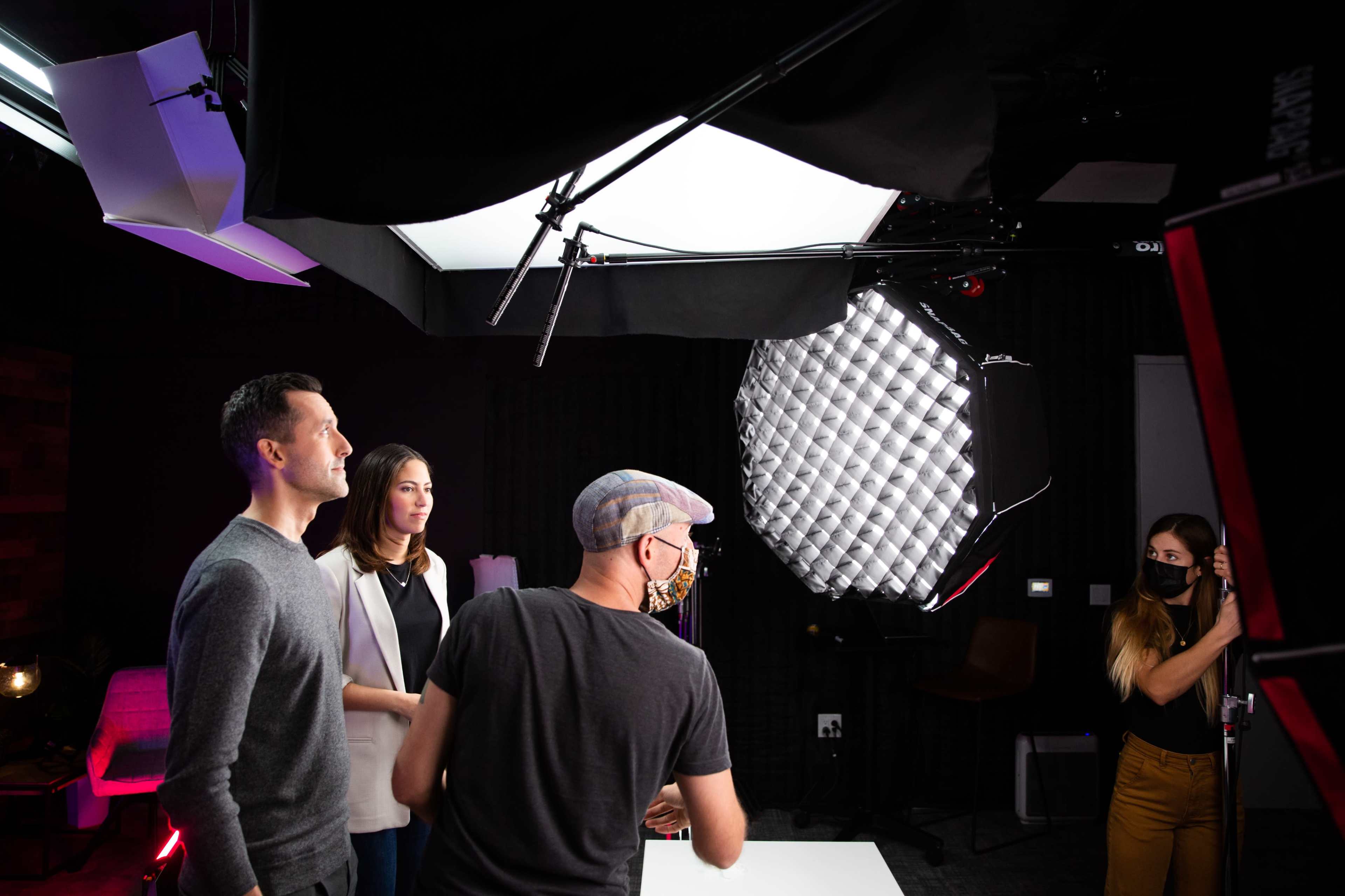 Four individuals are engaged in a professional setup for a video shoot, with bright lighting equipment and a camera positioned in front of them.