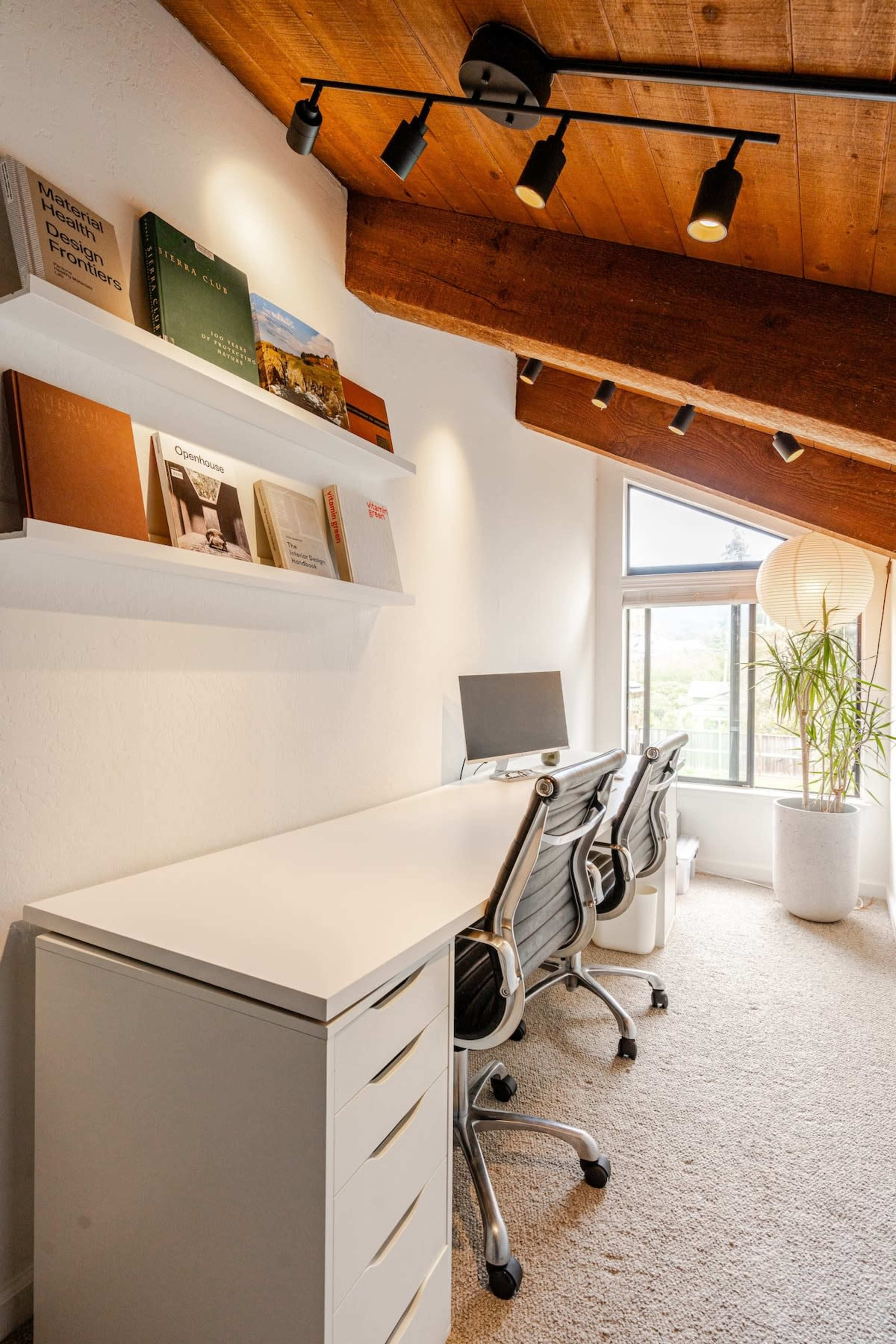The image shows a modern office space with a white desk, two ergonomic chairs, a computer, books displayed on a shelf, and a large window letting in natural light.