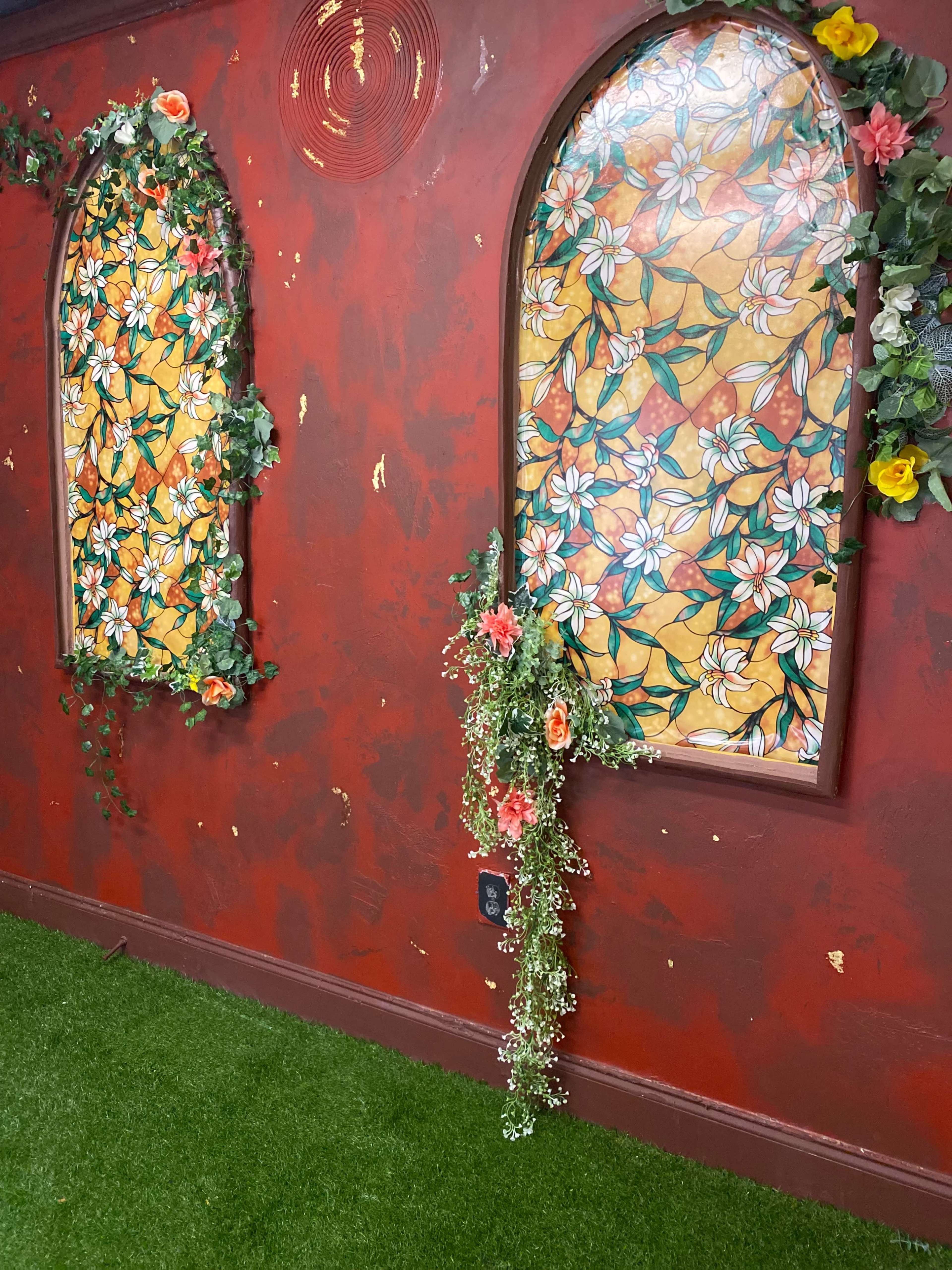 The image shows two arched windows with colorful stained glass, surrounded by floral decorations on a red wall, against a backdrop of green artificial grass.