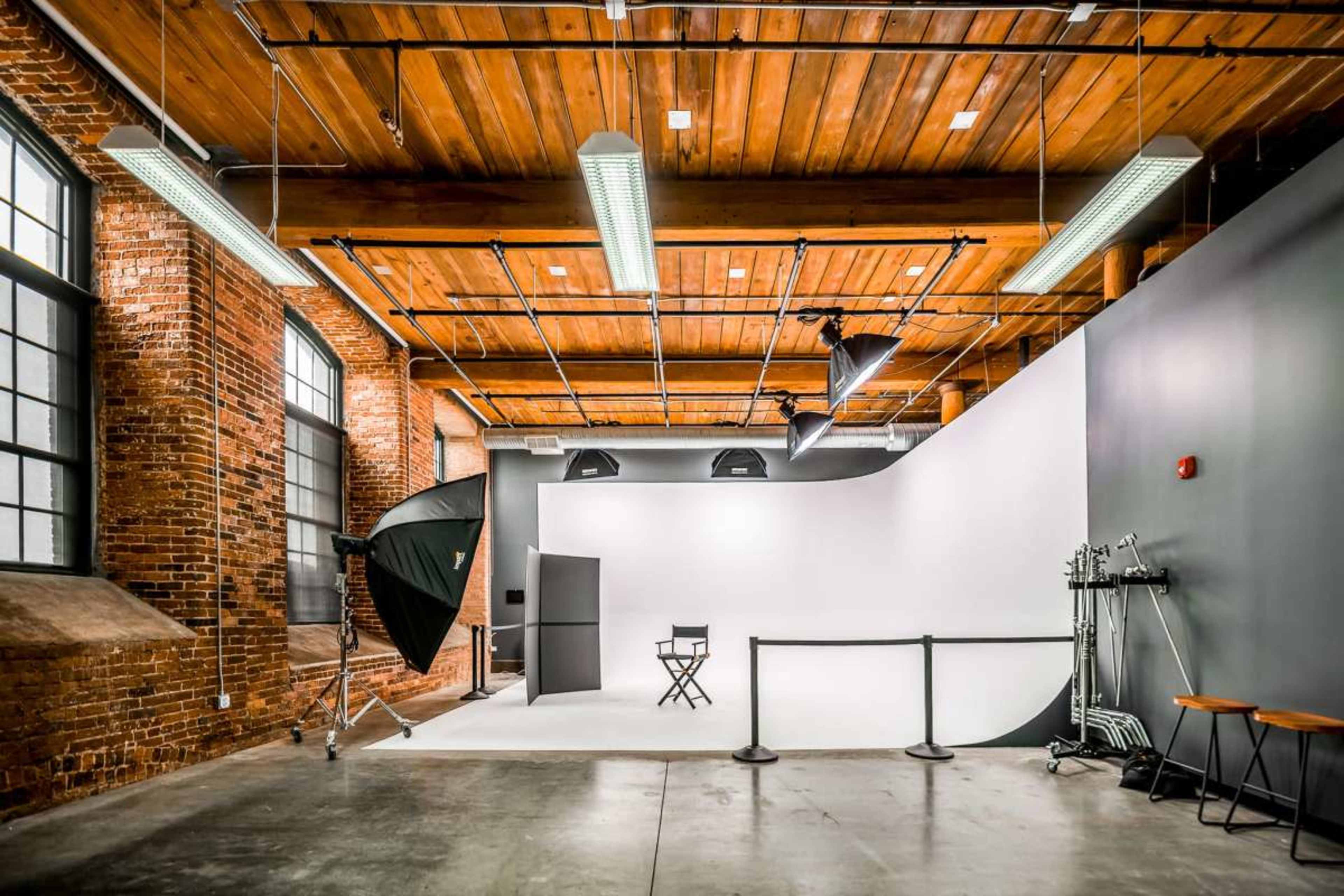 The image shows a spacious photo studio with a white backdrop, a director's chair, and various photography equipment against brick and wooden walls.