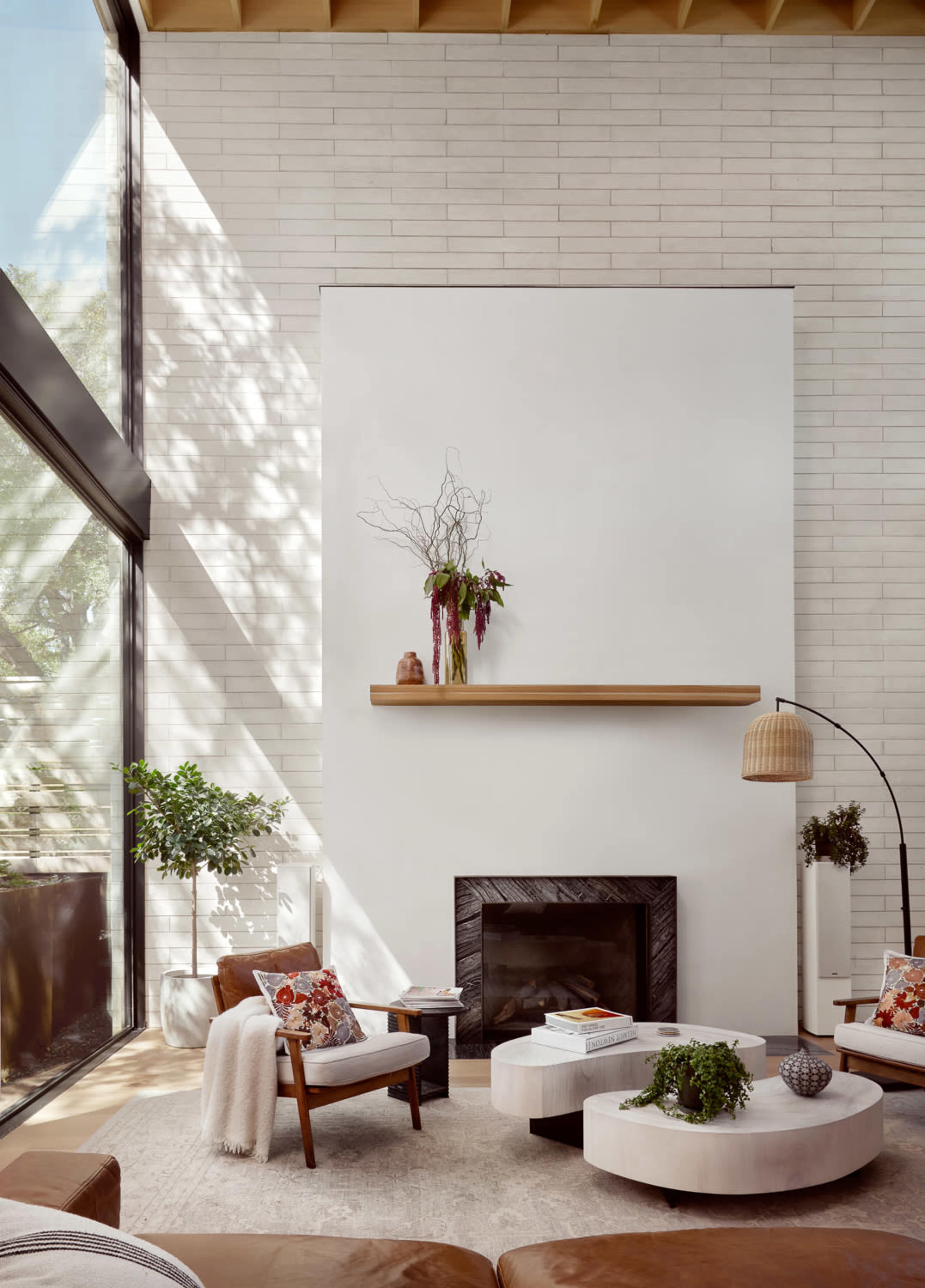 The scene depicts a modern living room with a white brick wall, a minimalist fireplace, comfortable seating, and large windows allowing natural light to enter.