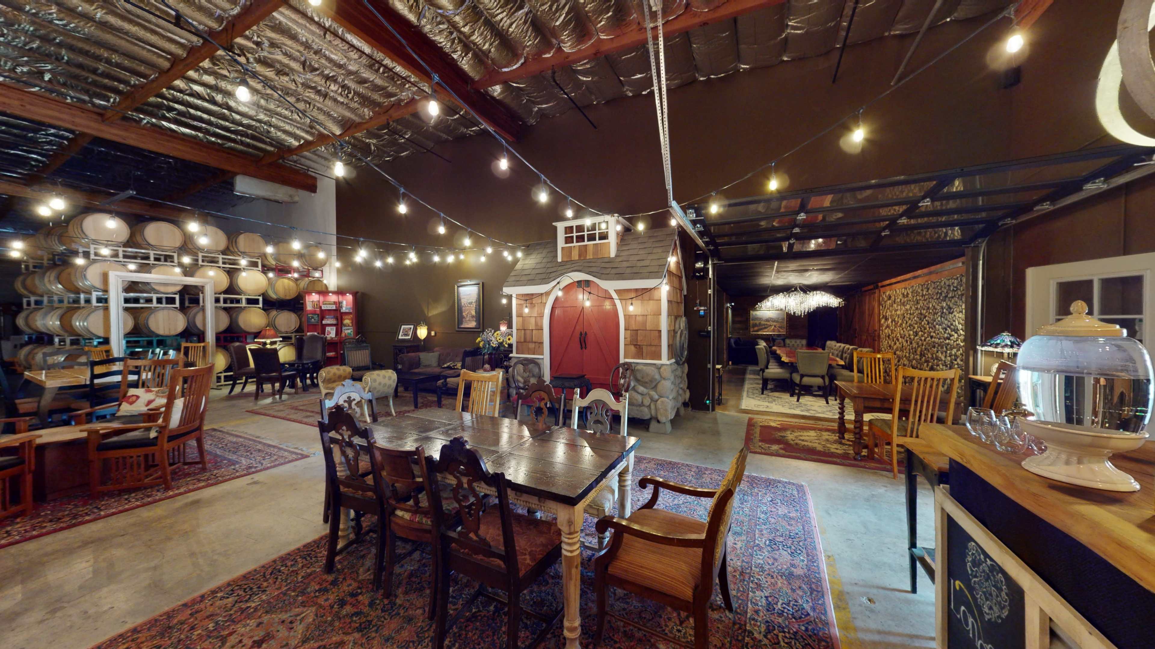 An indoor event space featuring a dining area with wooden tables and chairs, a decorative playhouse, wine barrels on display, and string lights overhead.