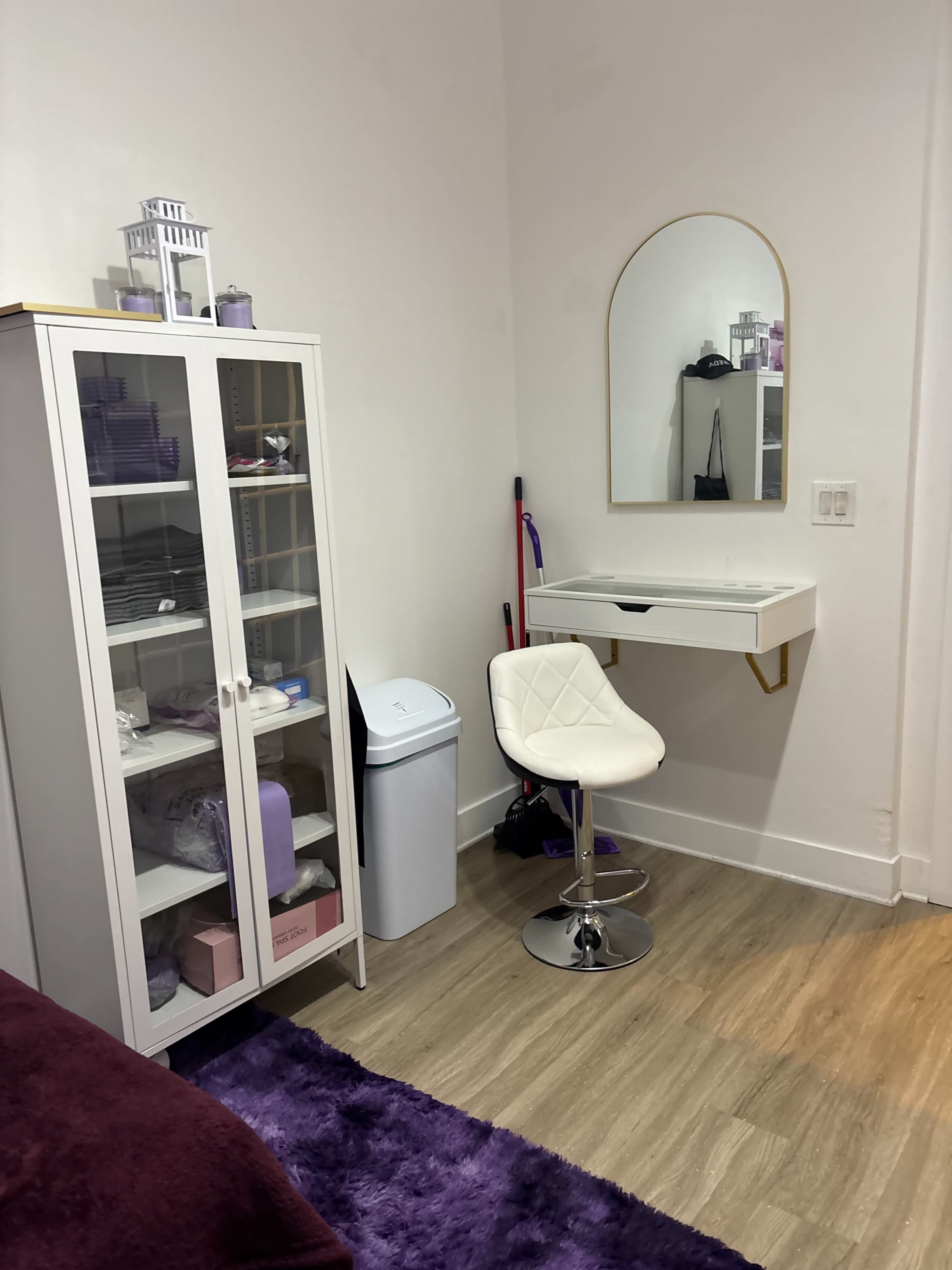 The image shows a small room with a white cabinet, a mirror hung on the wall, a desk with a chair, and a trash bin.