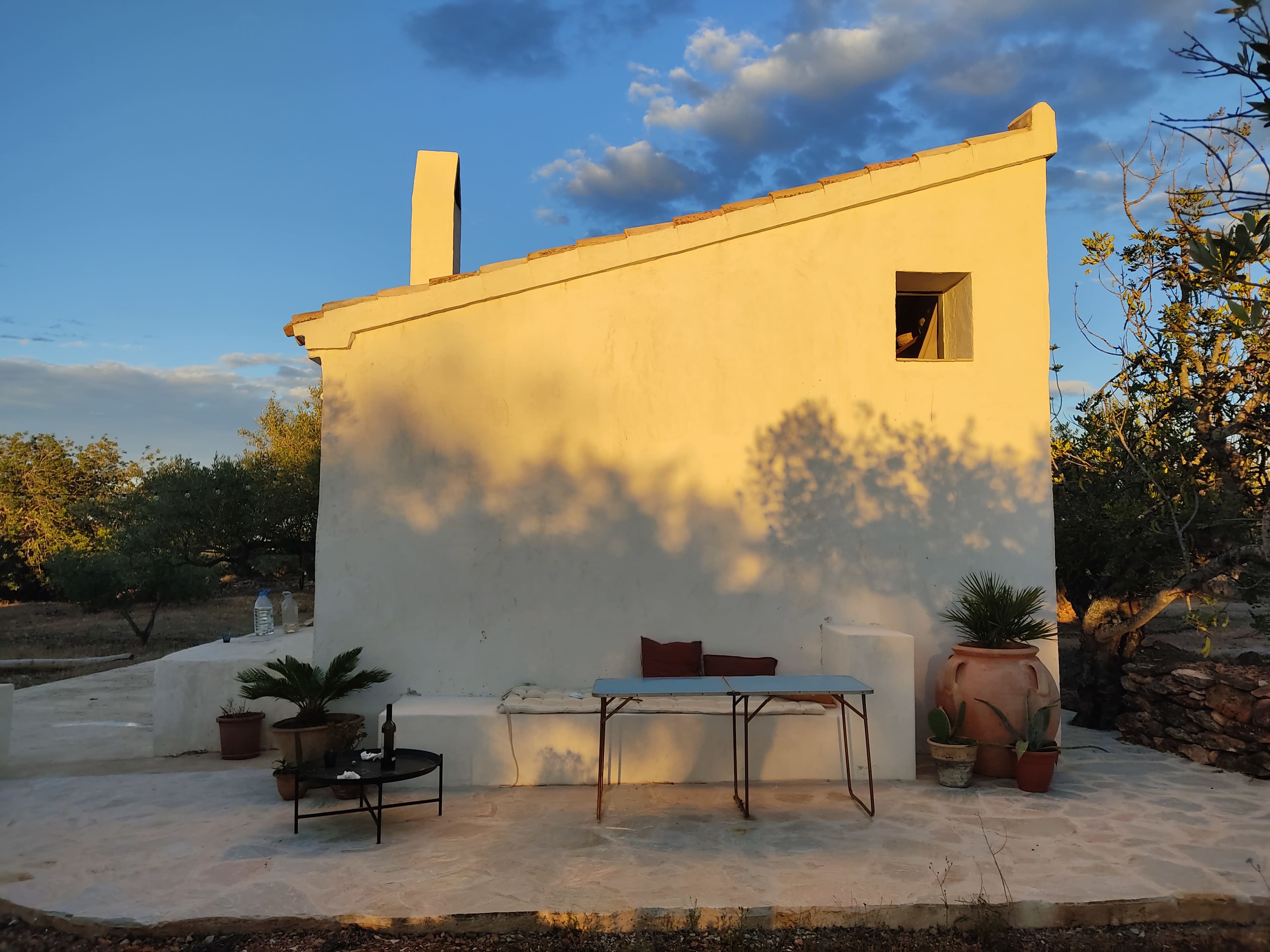 A simple white cottage with a slanted roof and a small window is set against a blue sky, accompanied by a table and potted plants on a stone patio.