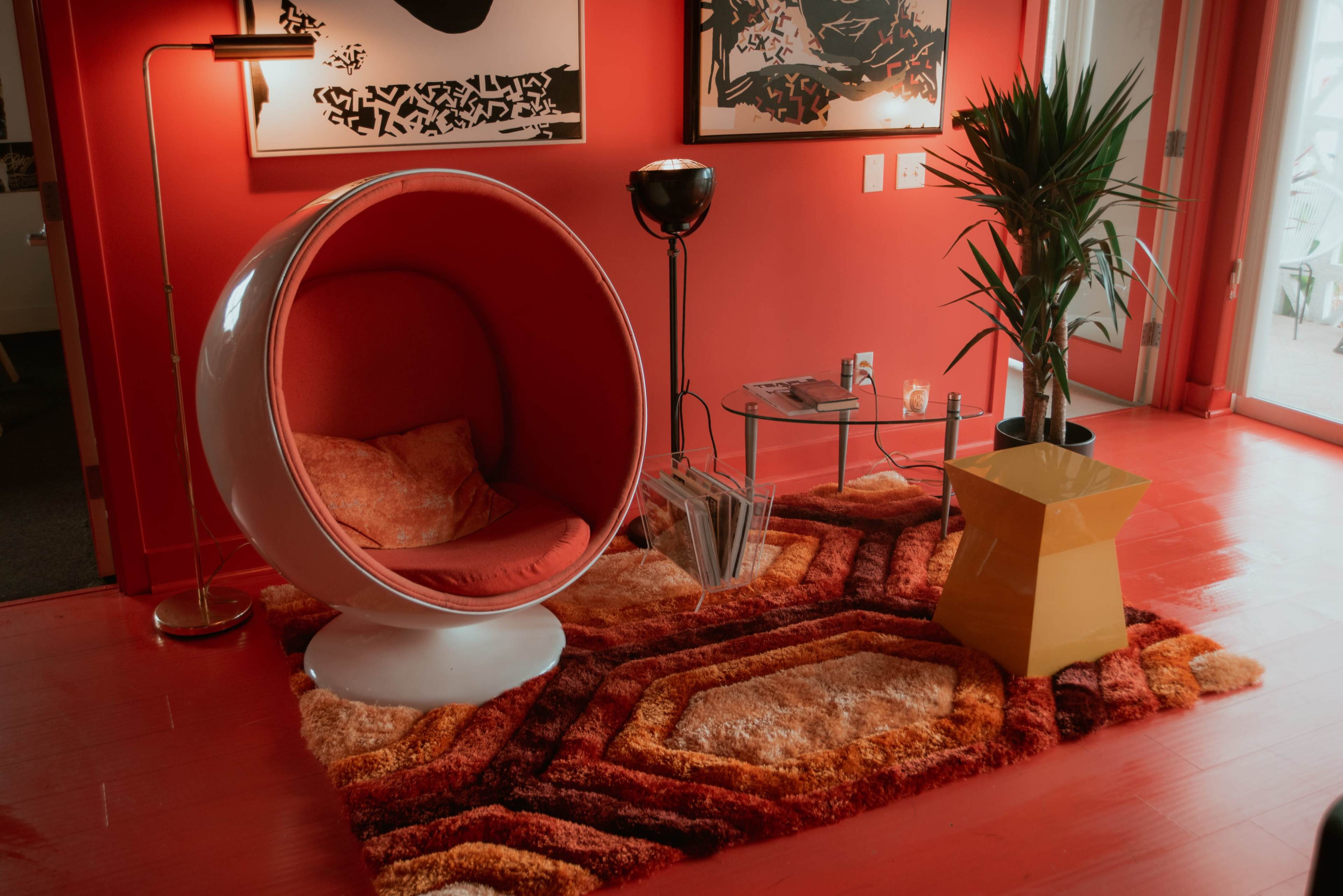 A modern red-themed living space features a round, white chair, a black lamp, a green plant, and a geometric yellow table on a colorful, textured rug.