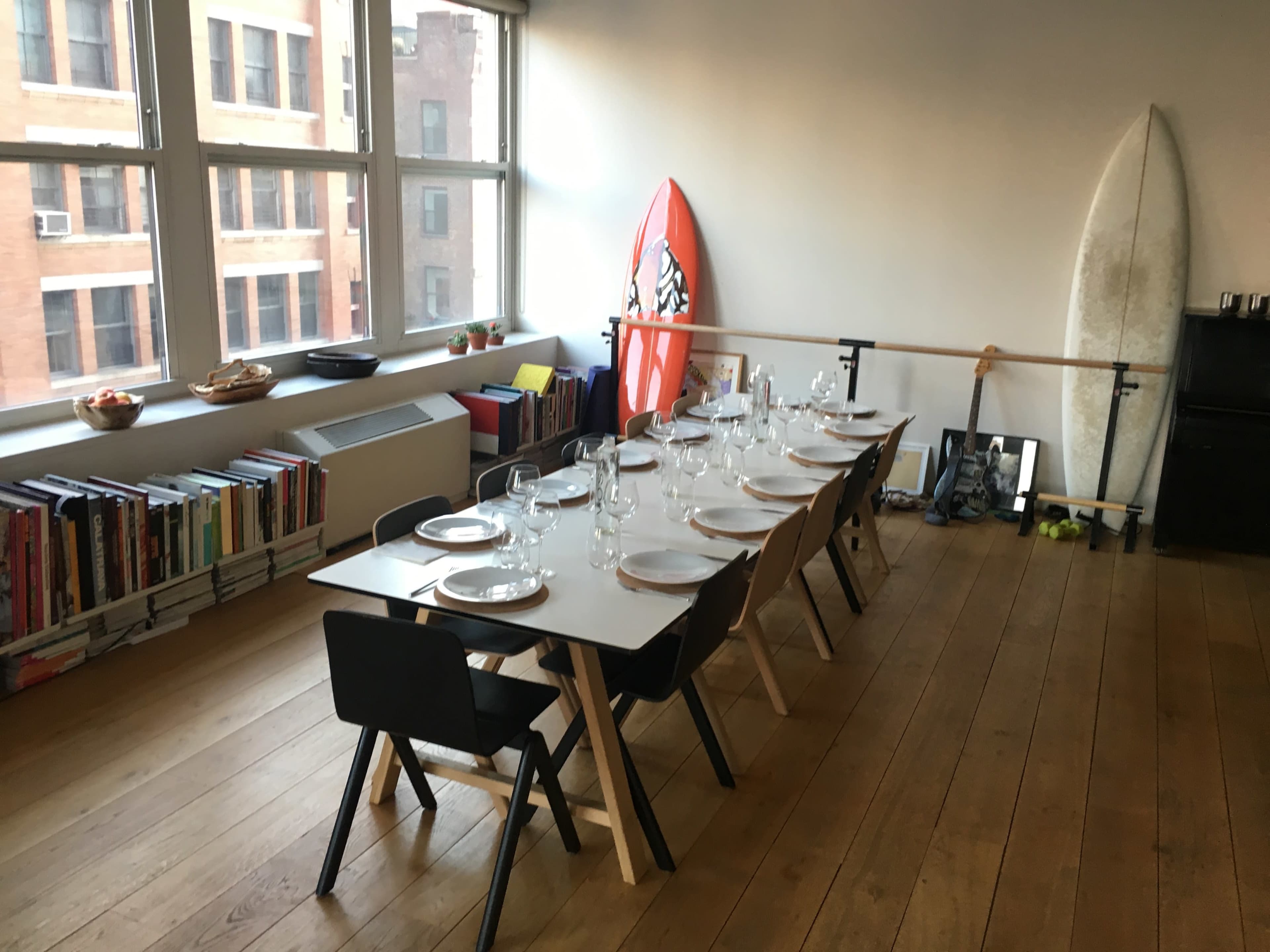A long, set dining table with dishes and glassware is positioned beside large windows, while surfboards lean against the wall in a spacious, wooden-floored room.