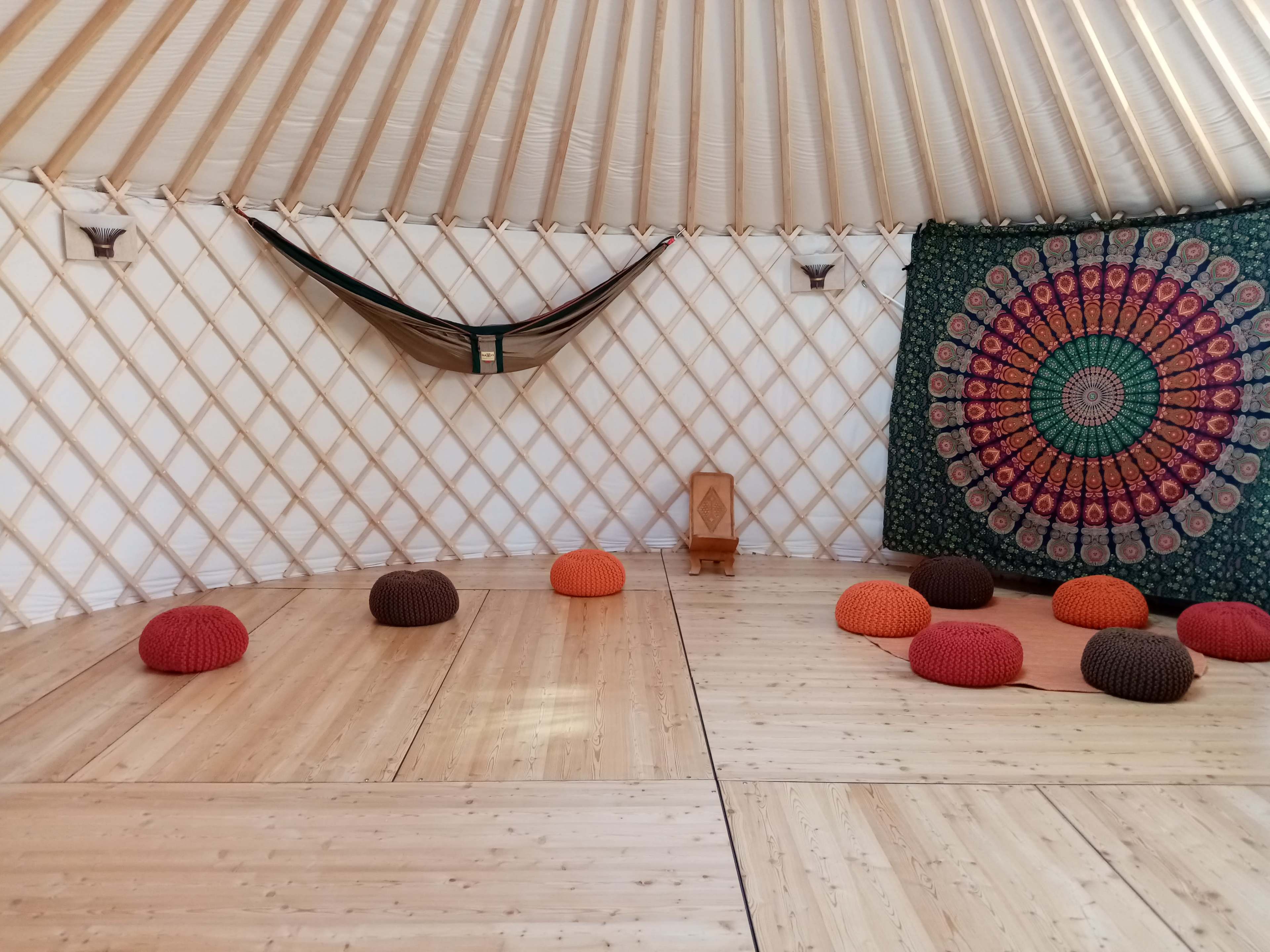 The interior of a yurt features a wooden floor, colorful round cushions arranged on the floor, a hammock hanging from the ceiling, and a patterned tapestry on one wall.