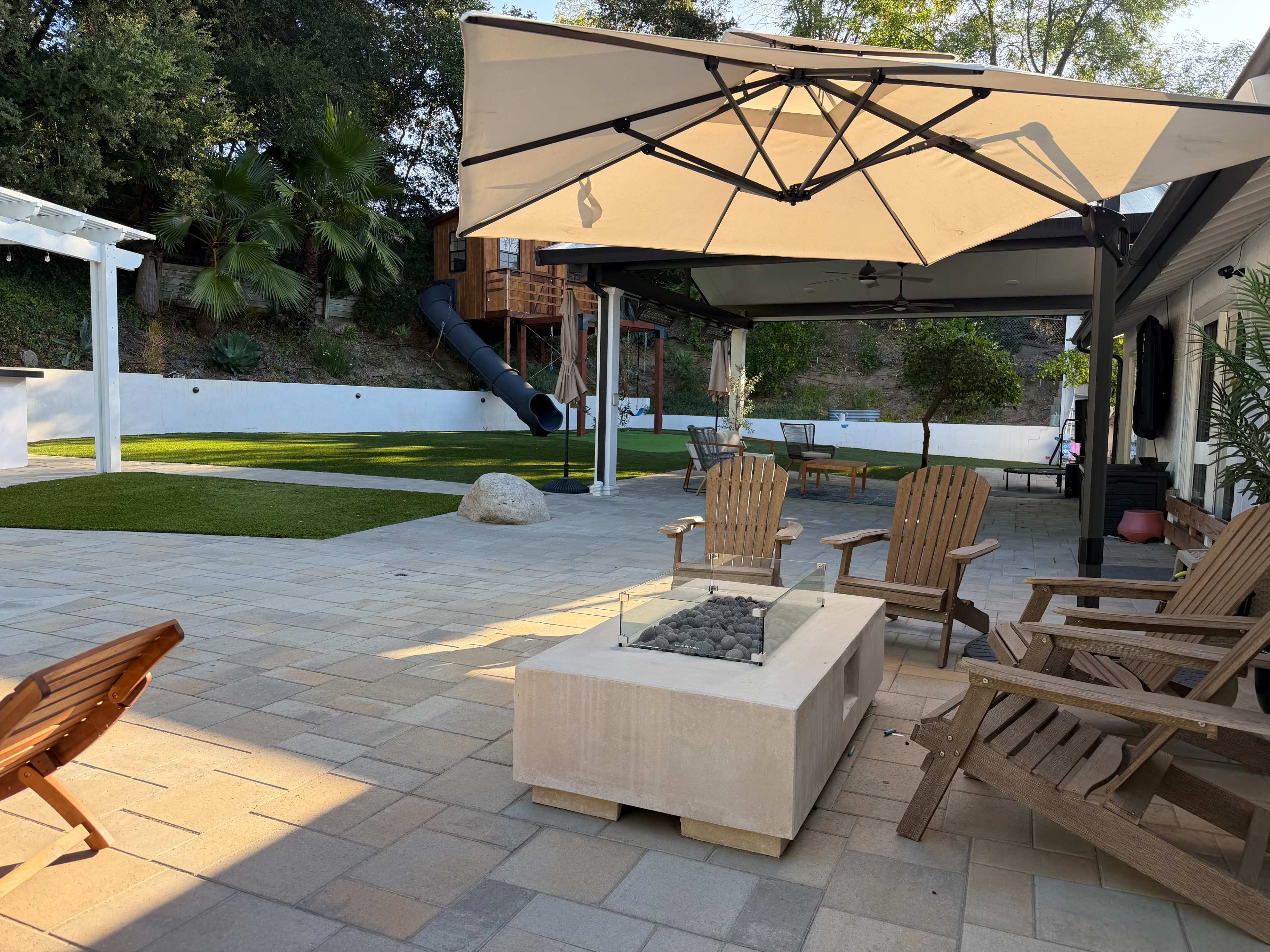 A backyard patio features a fire pit surrounded by wooden chairs, an umbrella, and grassy areas with a children's play structure and slide in the background.