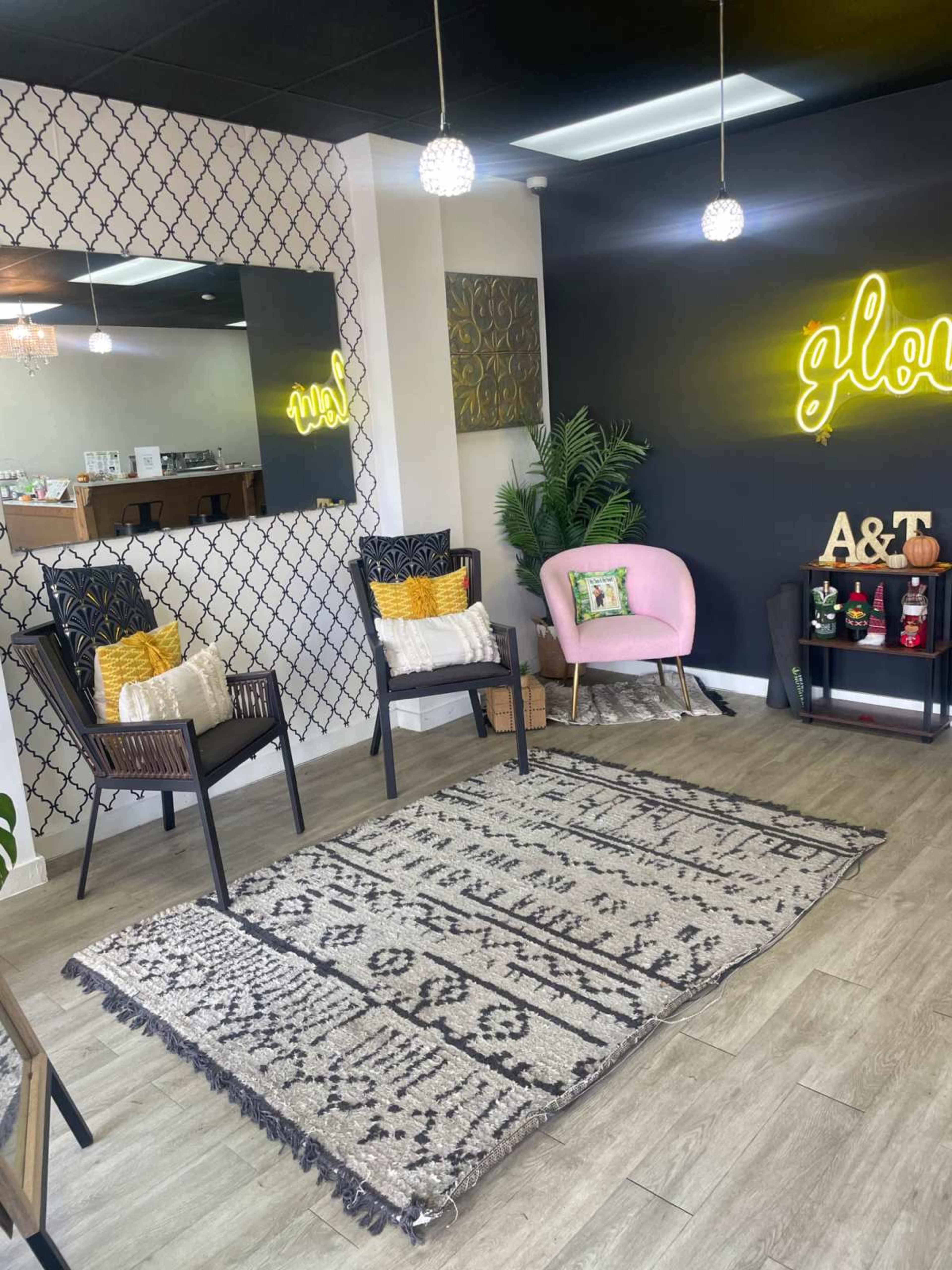 The image shows a cozy waiting area with two black chairs, a pink chair, a decorative rug, and a neon sign on a dark wall.