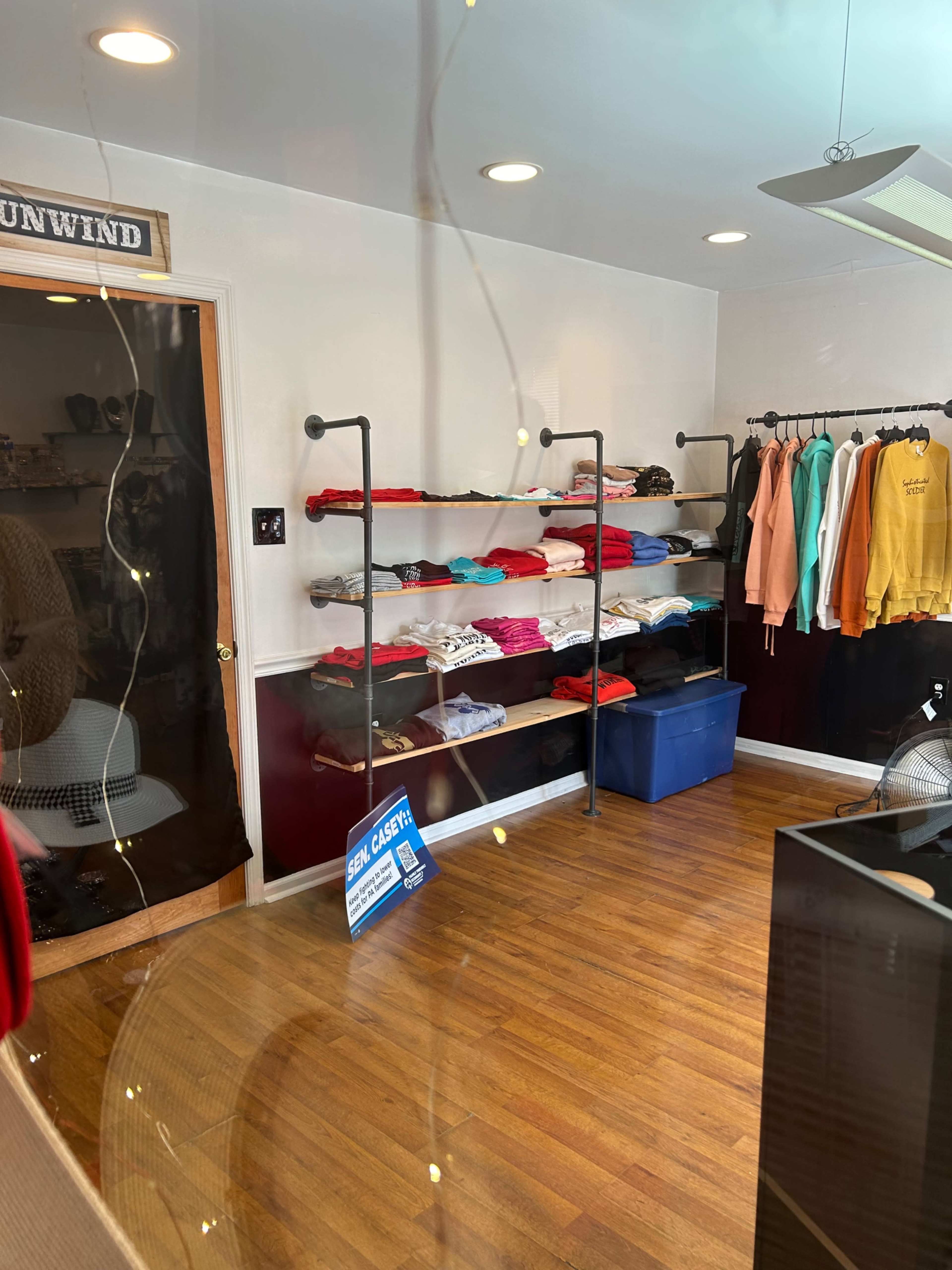 The image shows a clothing store interior featuring shelves of neatly folded shirts and hoodies in various colors, along with a blue storage bin and a fan.