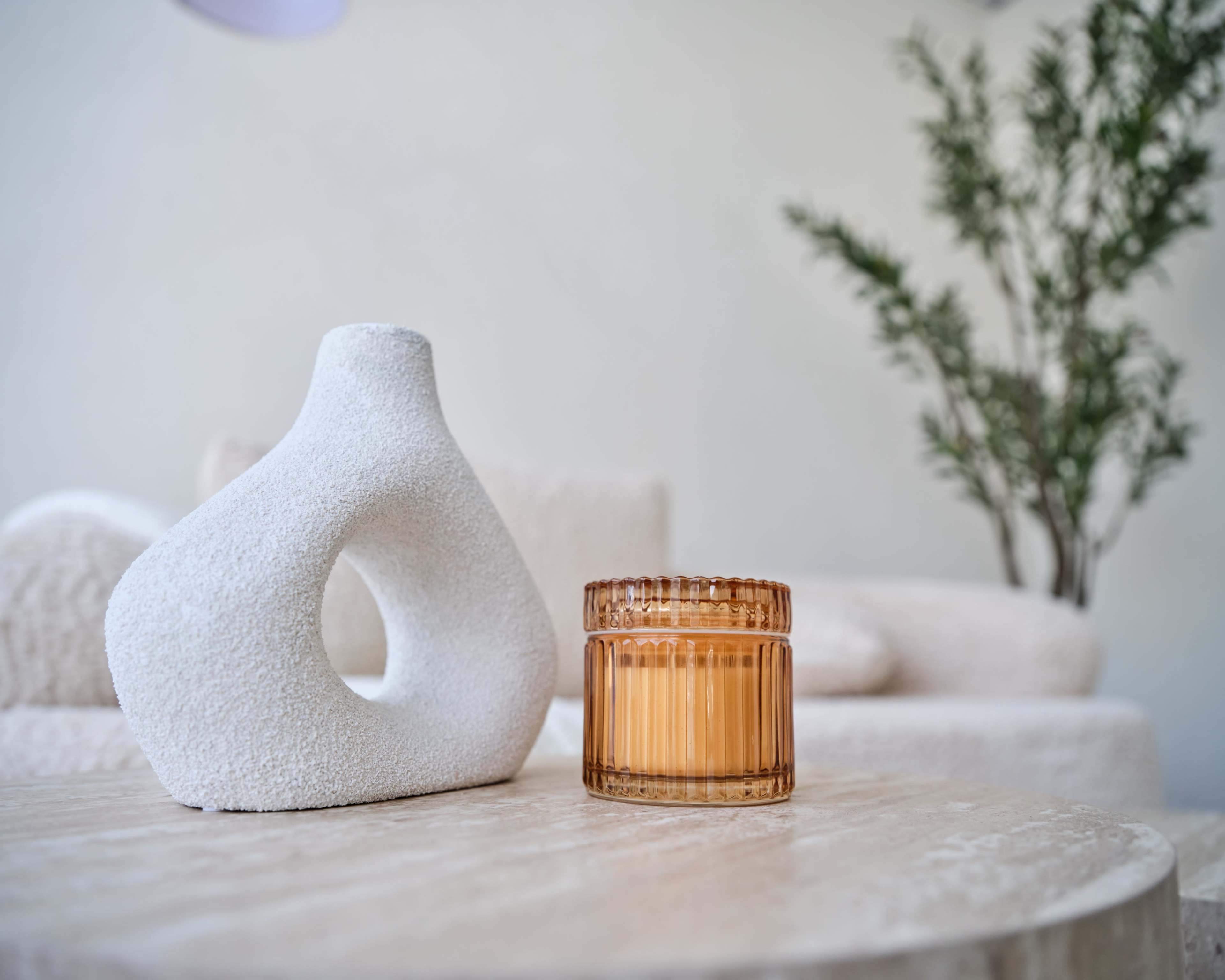 A textured white sculpture and a glass candle are placed on a marble table, with a green plant in the background.