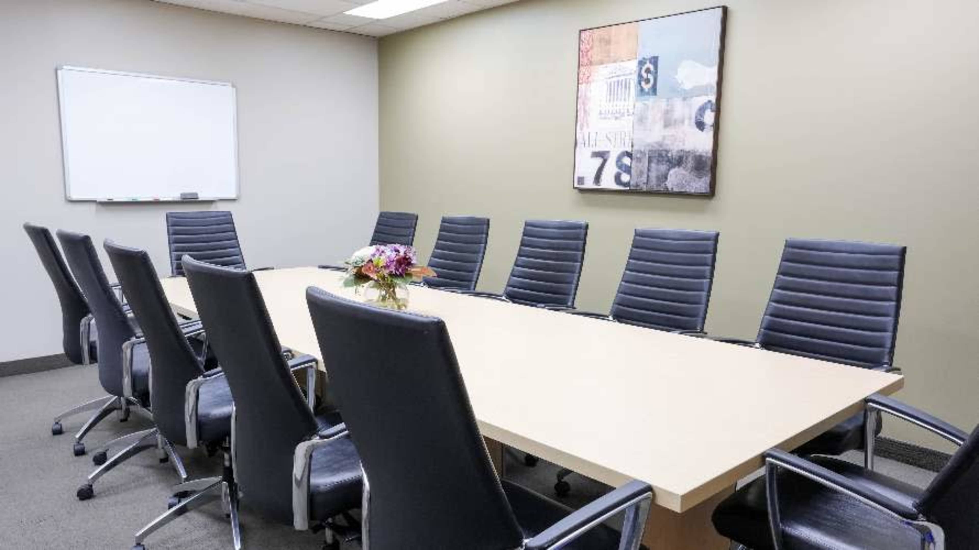 A conference room features a long table surrounded by eight black leather chairs and a floral centerpiece, with a whiteboard on one wall.