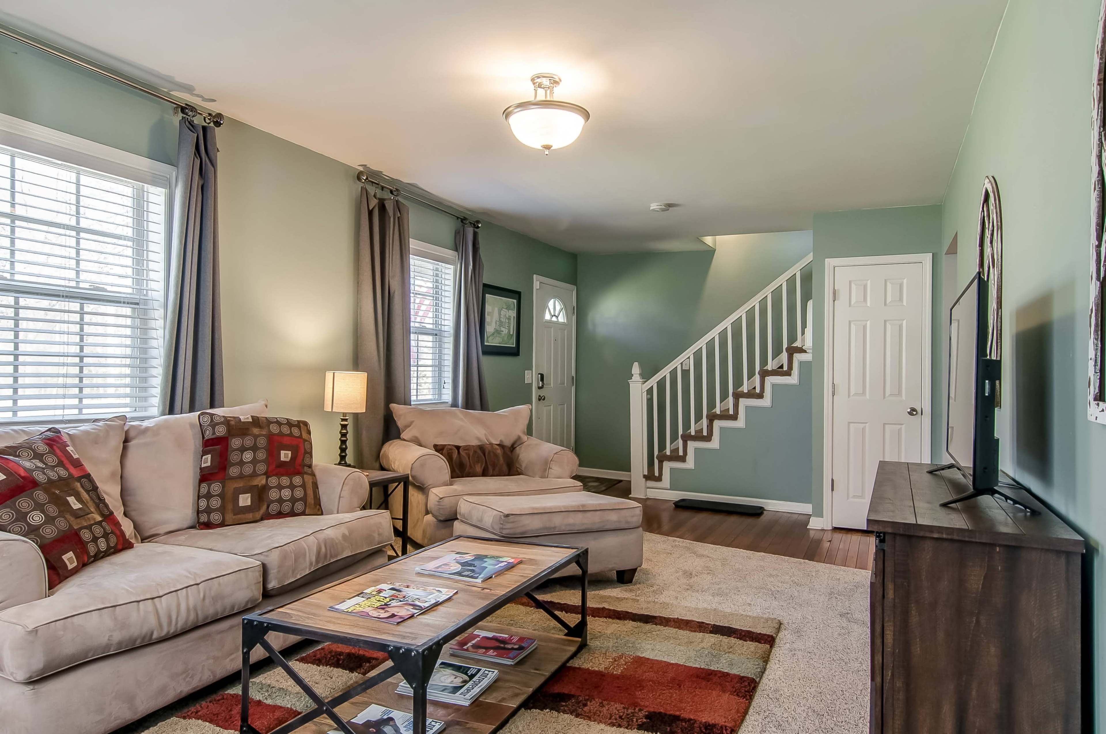 The living room features a beige sofa and armchair, a coffee table with magazines, and a staircase in the background.
