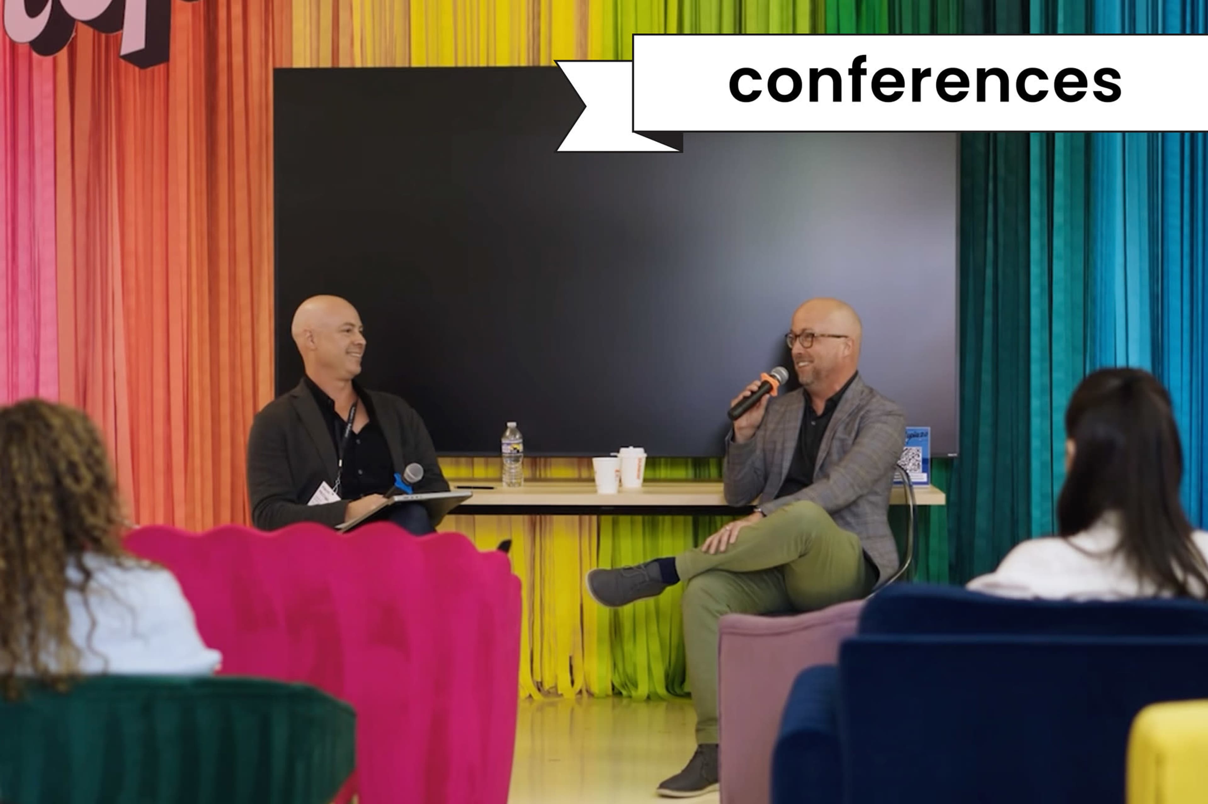 Two men are seated on colorful chairs in front of a large screen, engaging in a discussion during a conference setting.