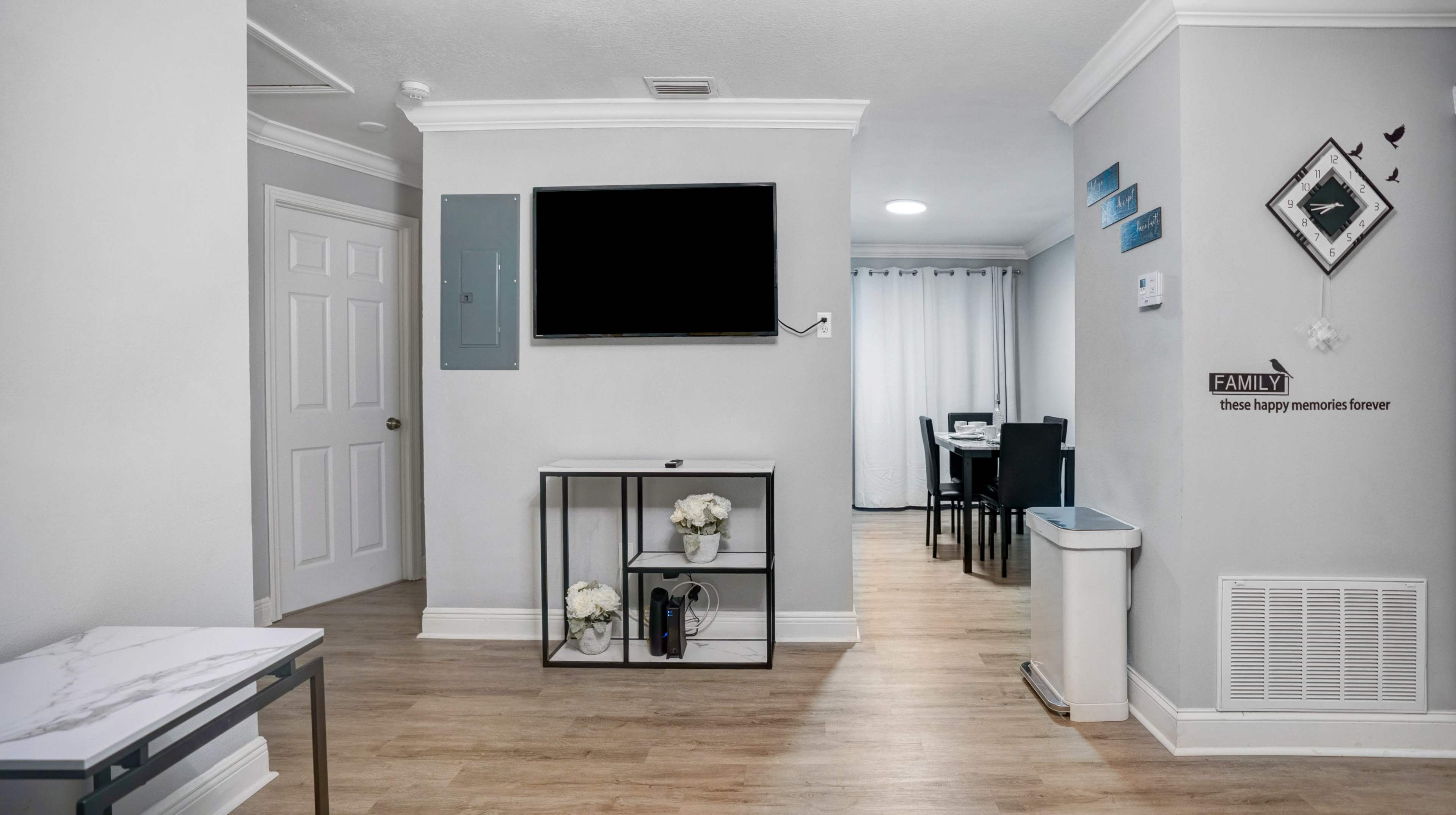 A modern living area with a wall-mounted TV, a small storage unit with decorative items, and a view into a dining area with a table and chairs.