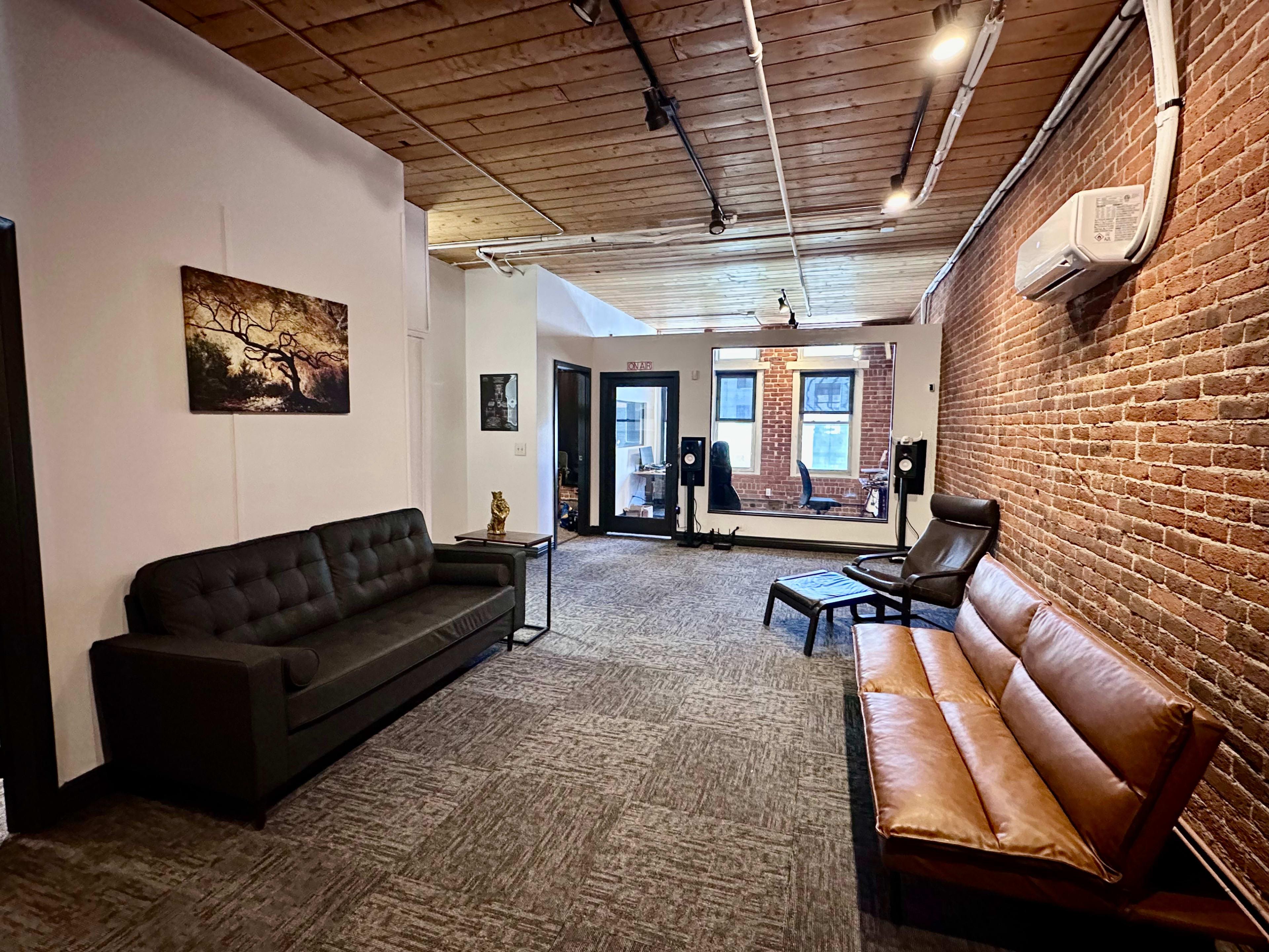 The image shows a modern interior space with a black sofa and a brown leather couch against a brick wall, and large windows in the background.