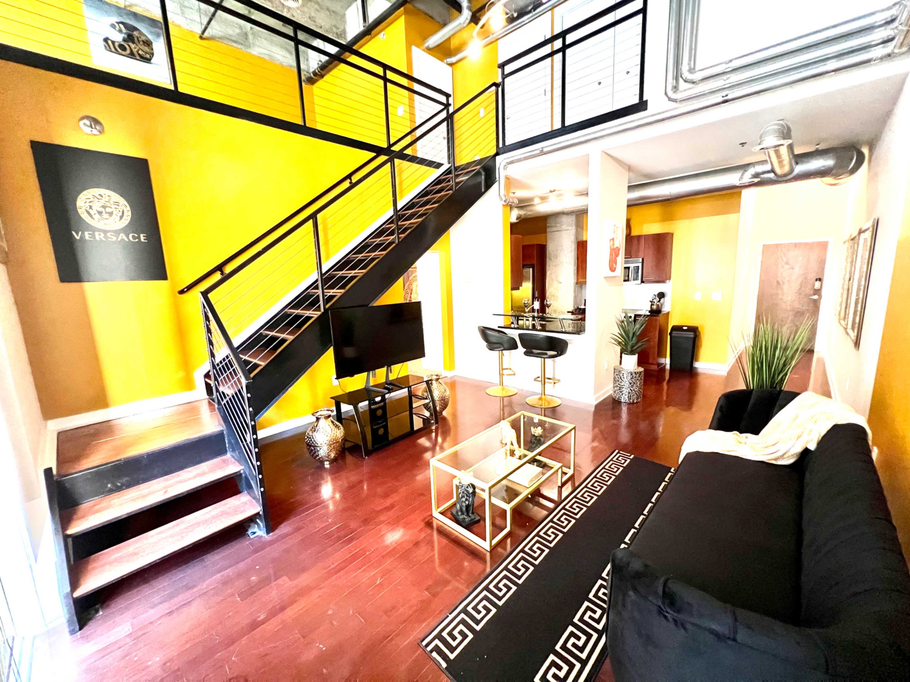 A modern loft living space featuring a black couch, a glass coffee table, a staircase leading to an upper area, and yellow walls adorned with a Versace poster.