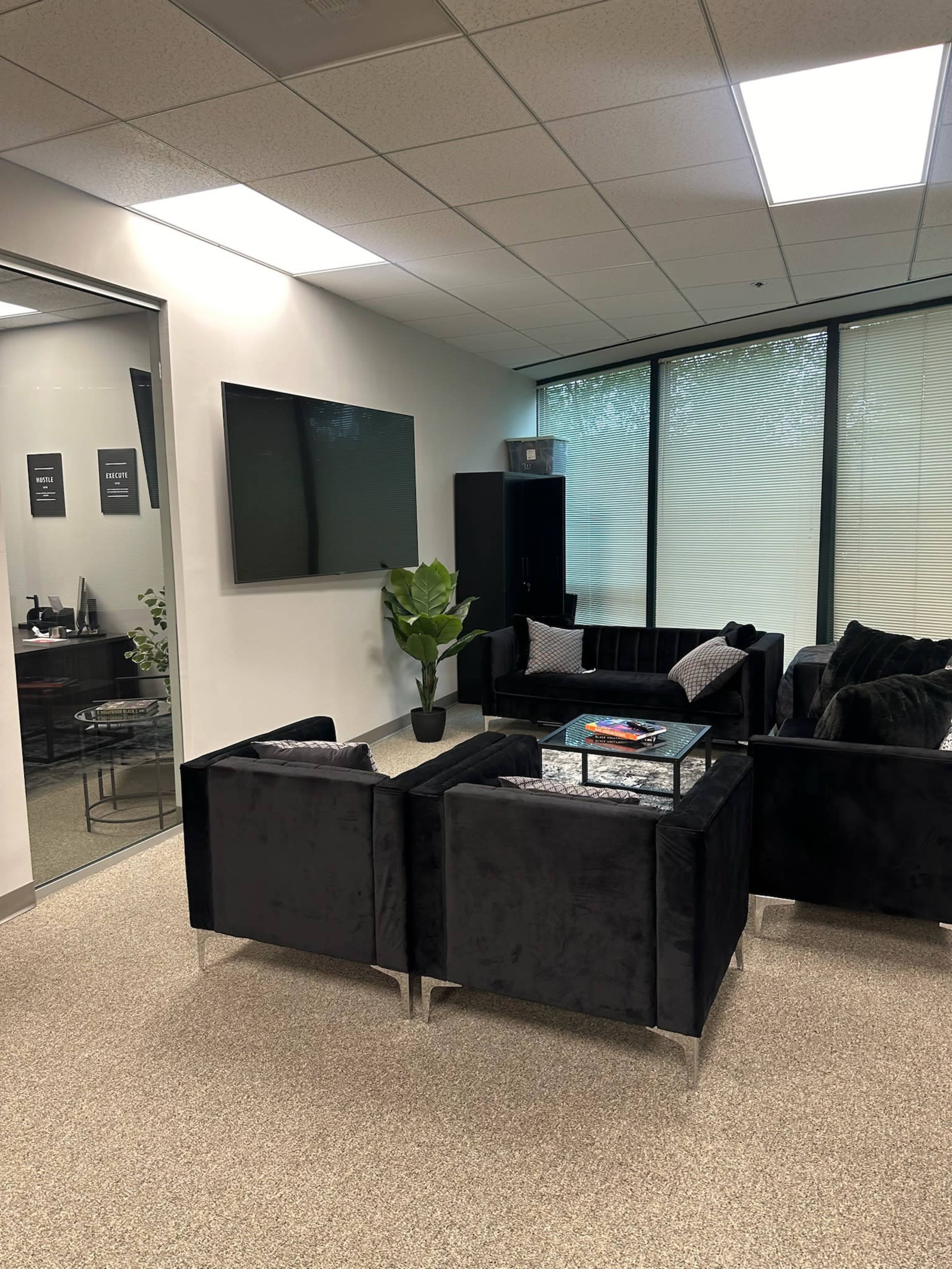 The image shows a modern office waiting area furnished with black velvet couches, a glass coffee table, and a potted plant, with large windows providing natural light.