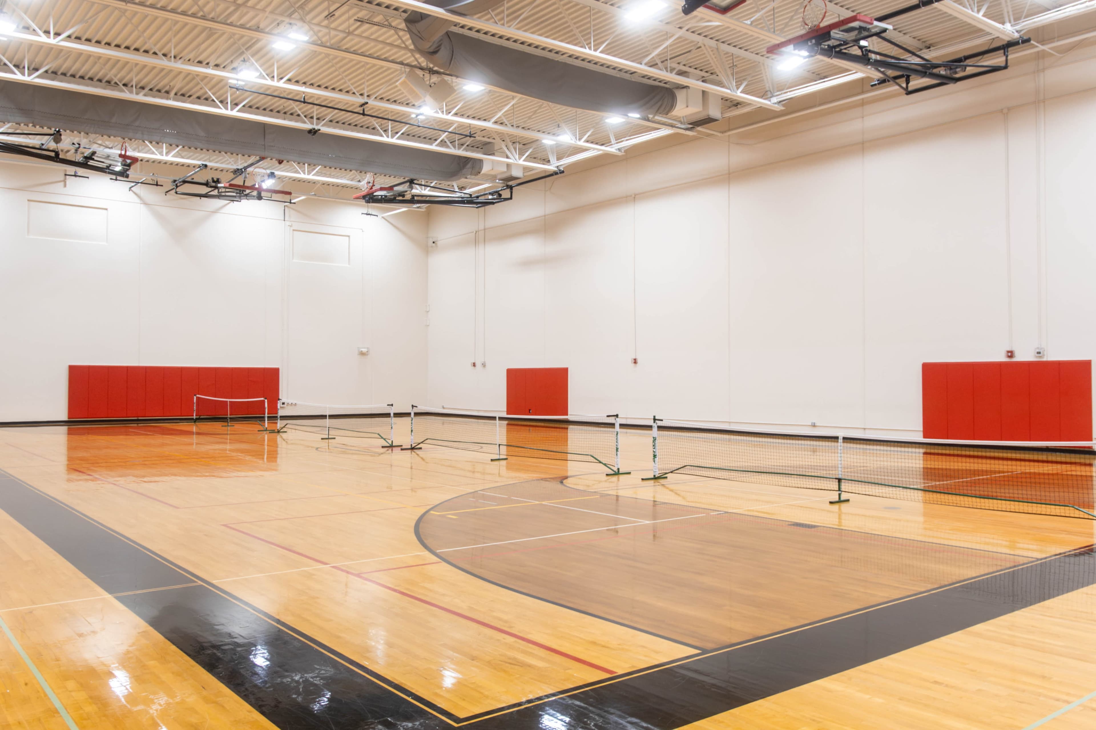 The image shows an empty indoor gymnasium with tennis nets set up on a polished wooden floor and red wall sections.