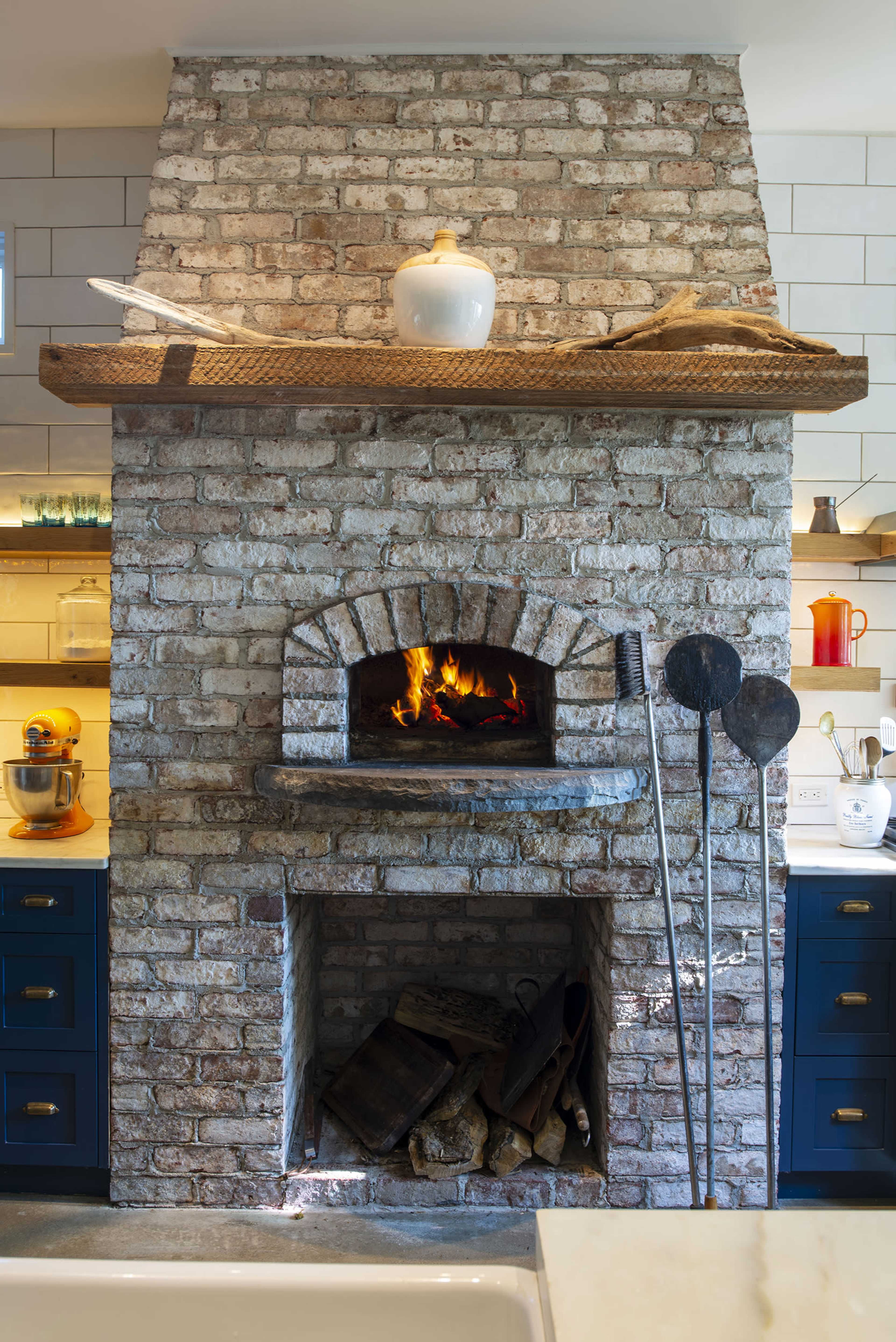 A stone fireplace with a wooden mantel is the focal point of a modern kitchen, surrounded by blue cabinets and a well-organized countertop.