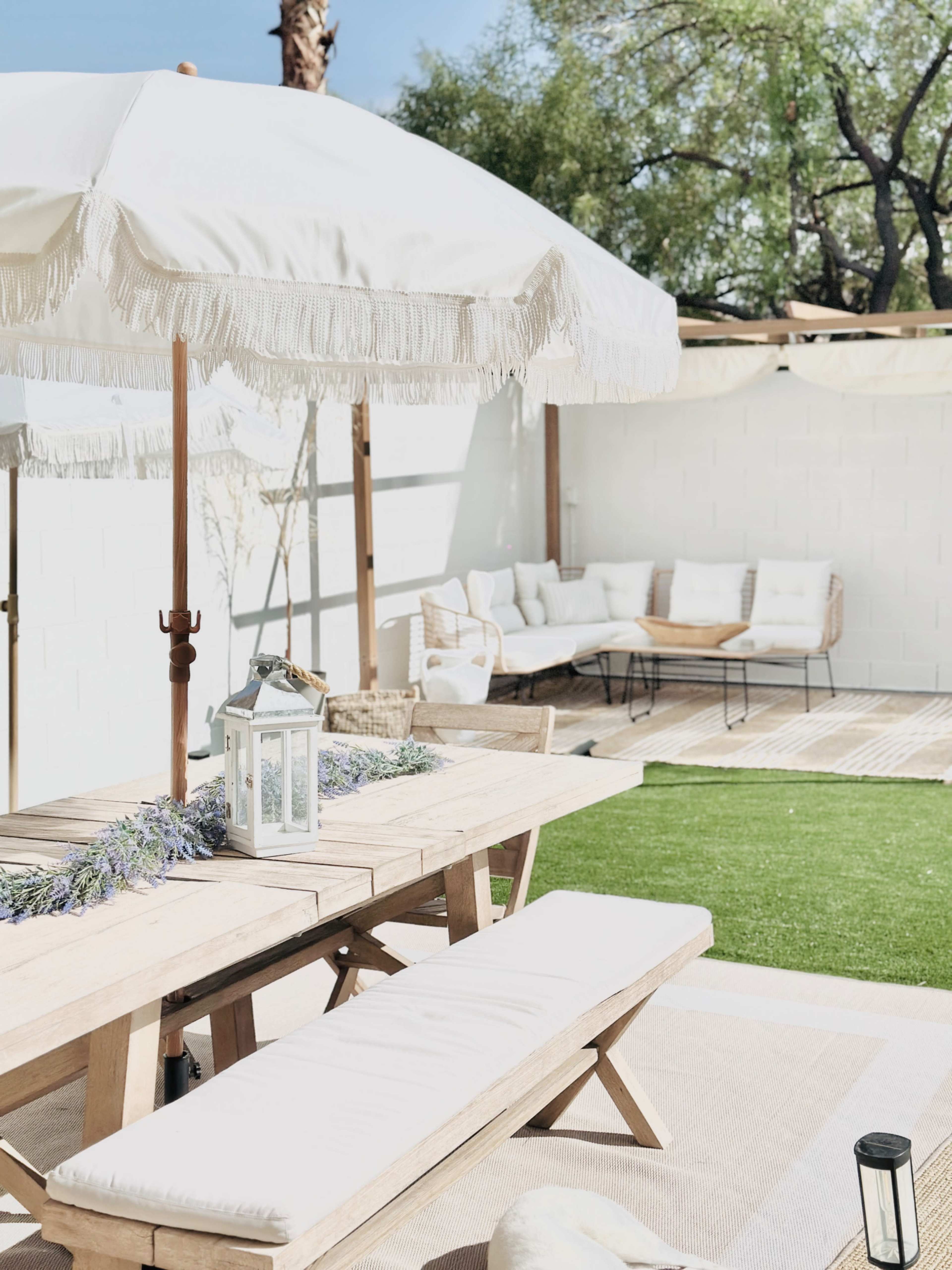 A wooden dining table with a bench is set under a white parasol in a landscaped outdoor area featuring a grass lawn and seating arrangement.