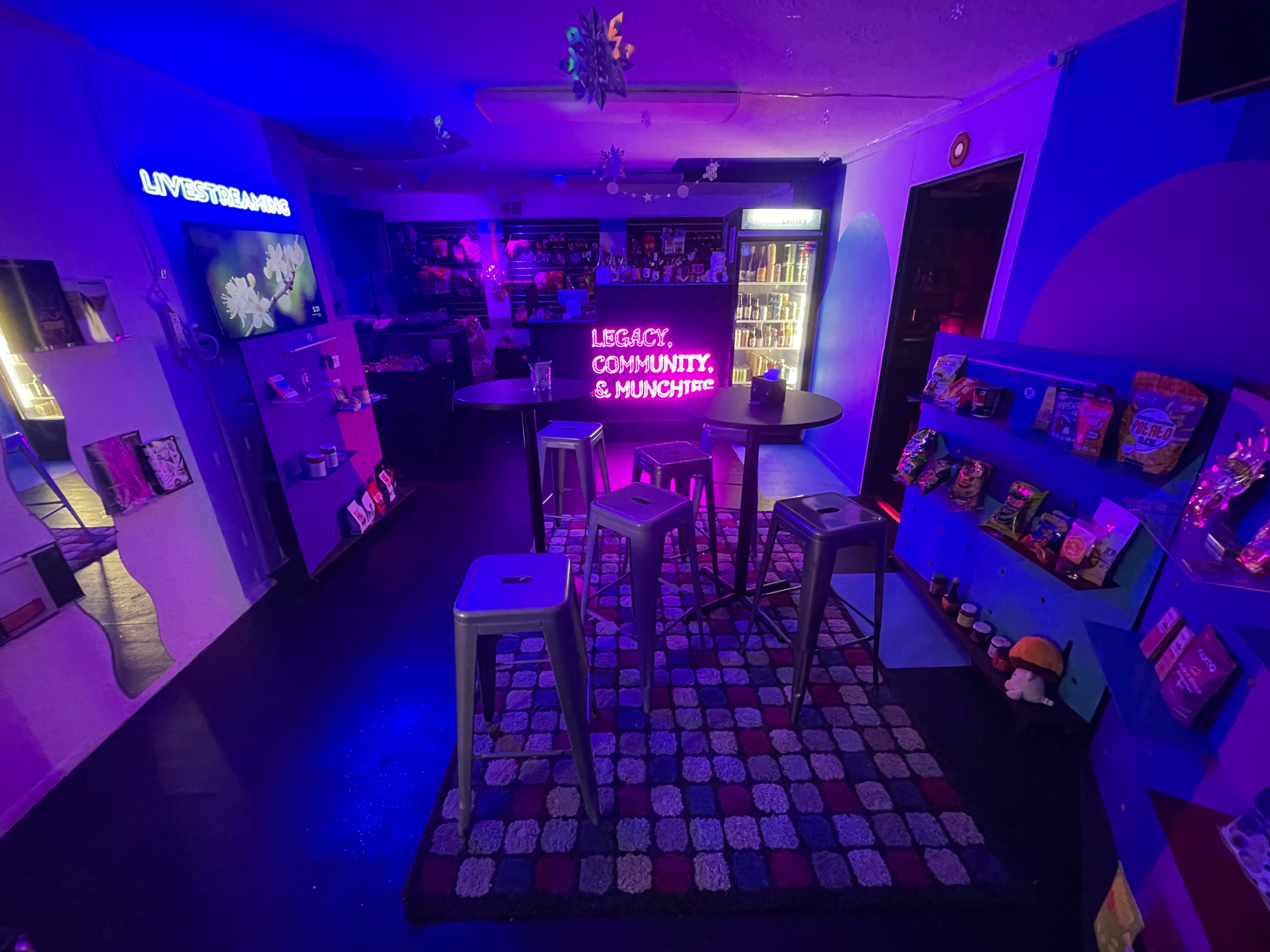 The image shows a colorful bar area with modern furniture, featuring neon signs and shelves filled with snacks.