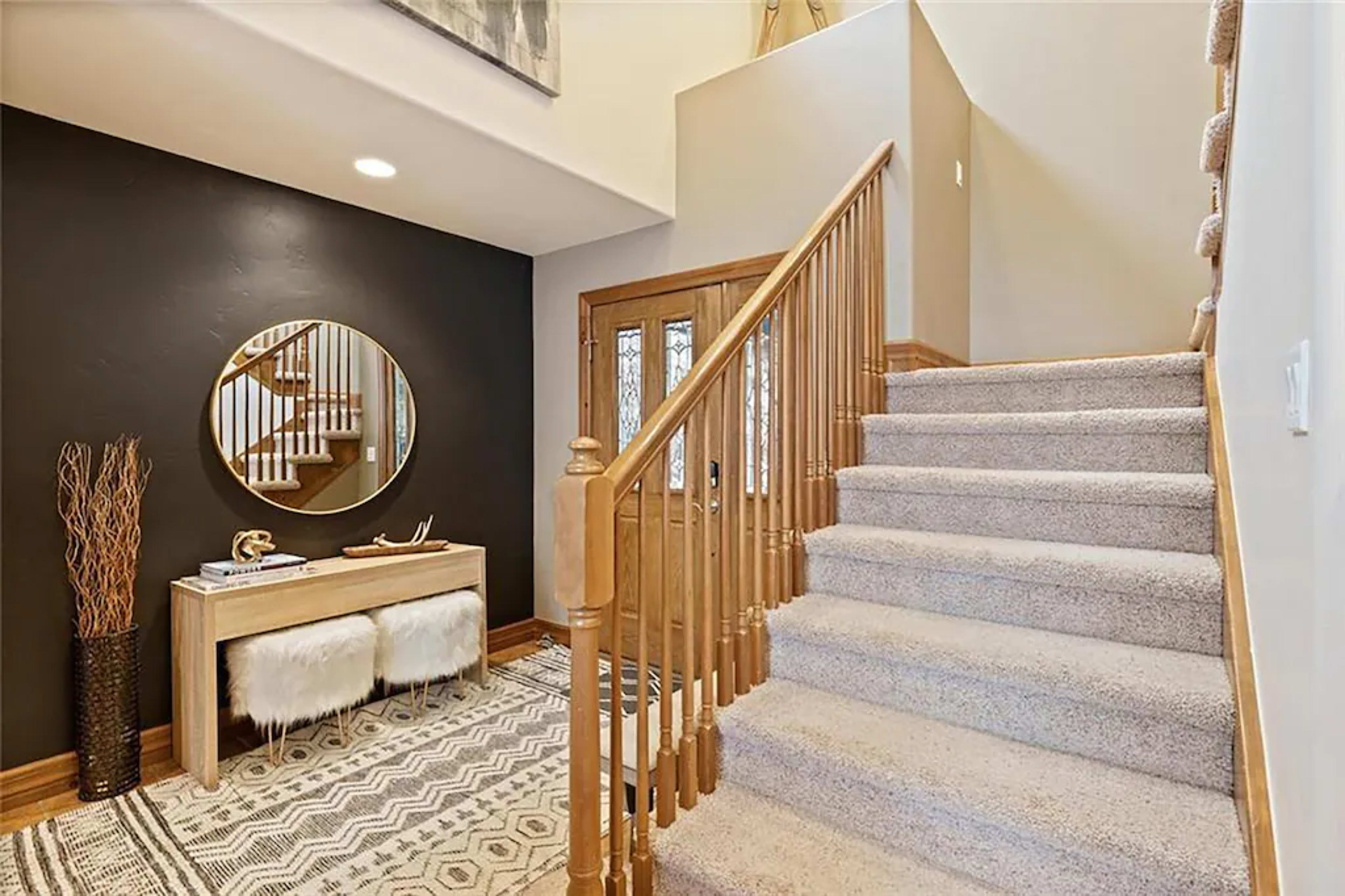 The image shows a staircase leading to an upper level, with a console table and a large round mirror positioned against a dark wall in the entryway.