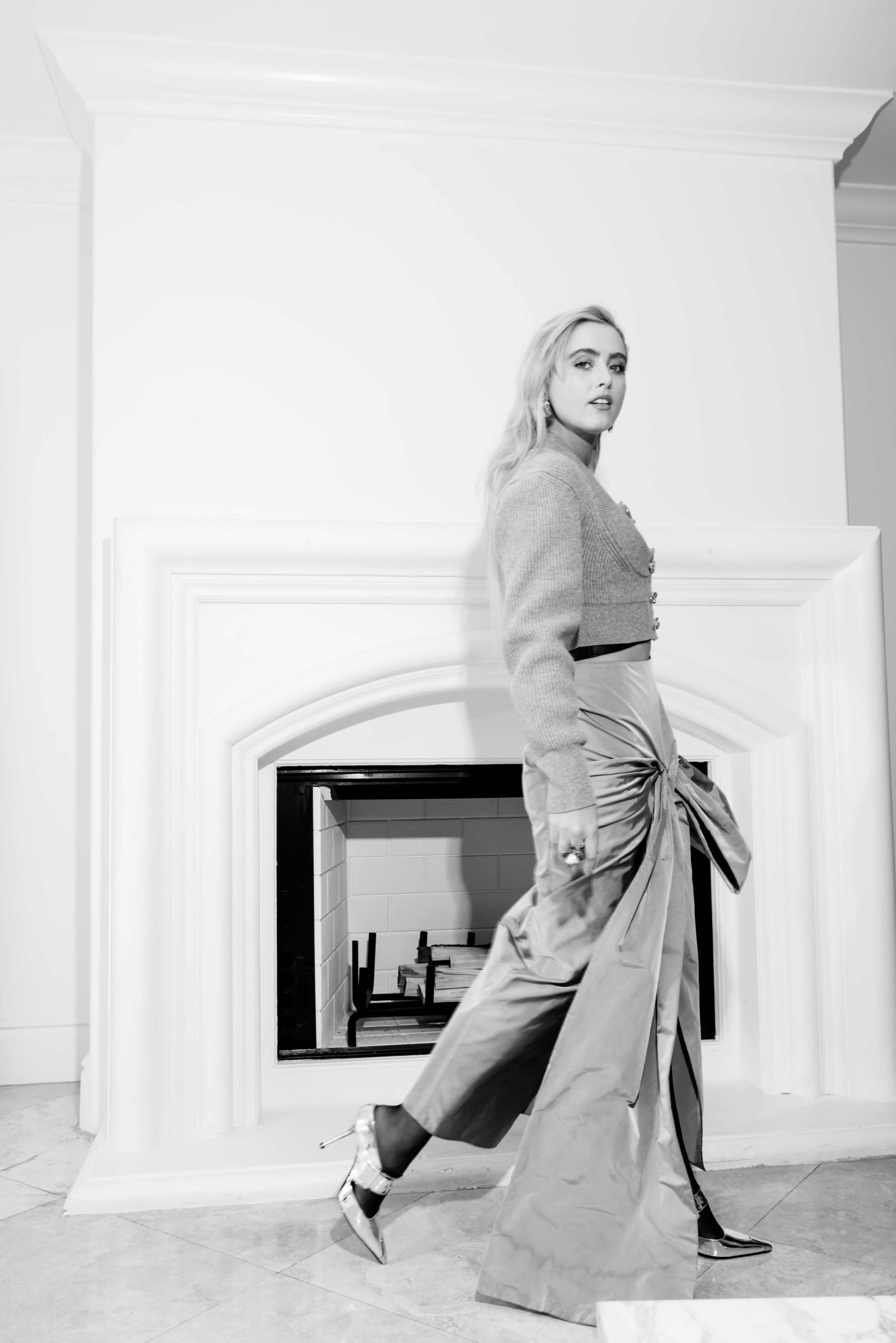 A person walks past a white fireplace while wearing a stylish outfit, featuring a textured top and a flowy skirt.