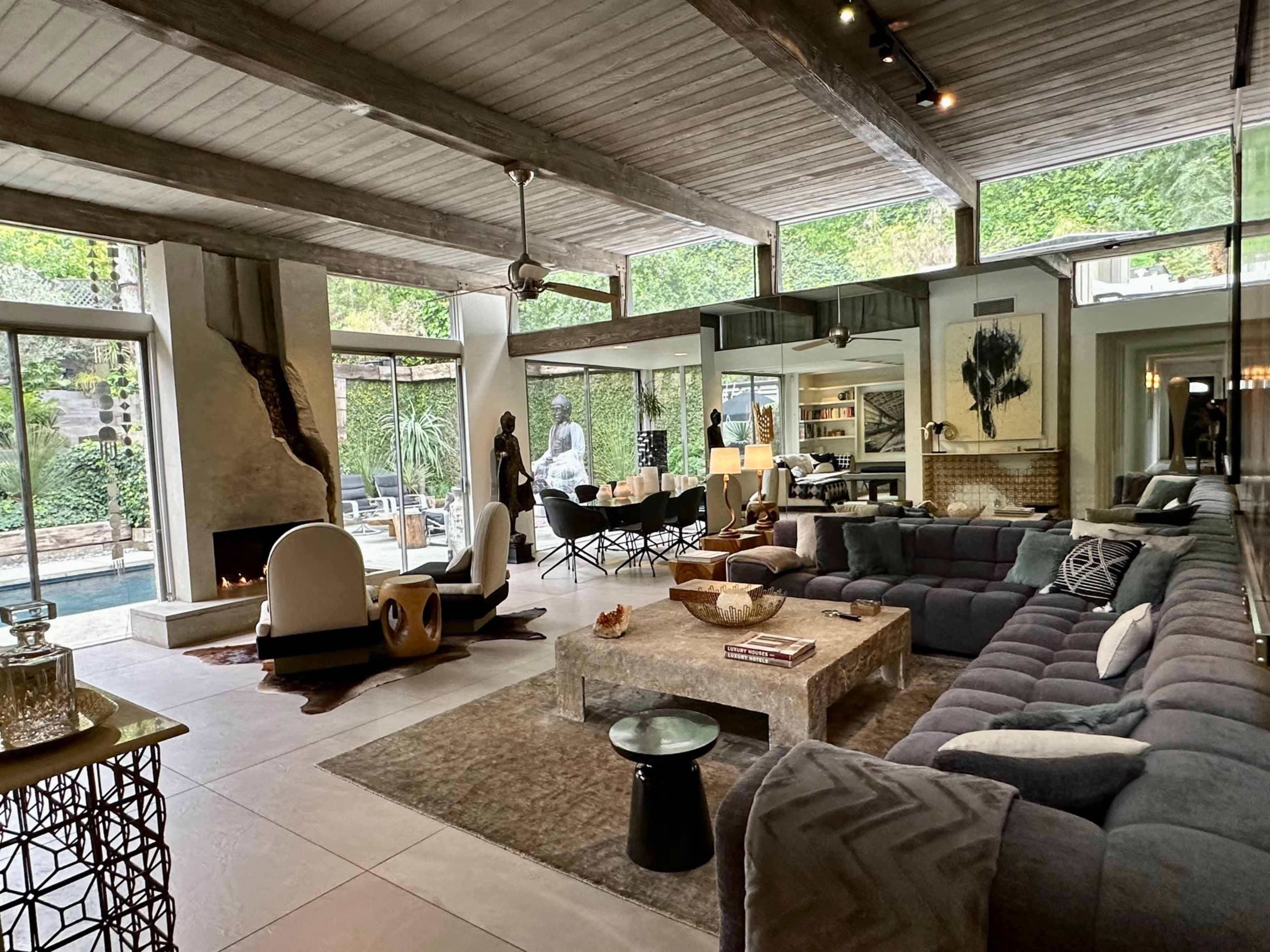 A modern living room featuring a large sectional sofa, a stone coffee table, and large windows overlooking a garden area with a pool.