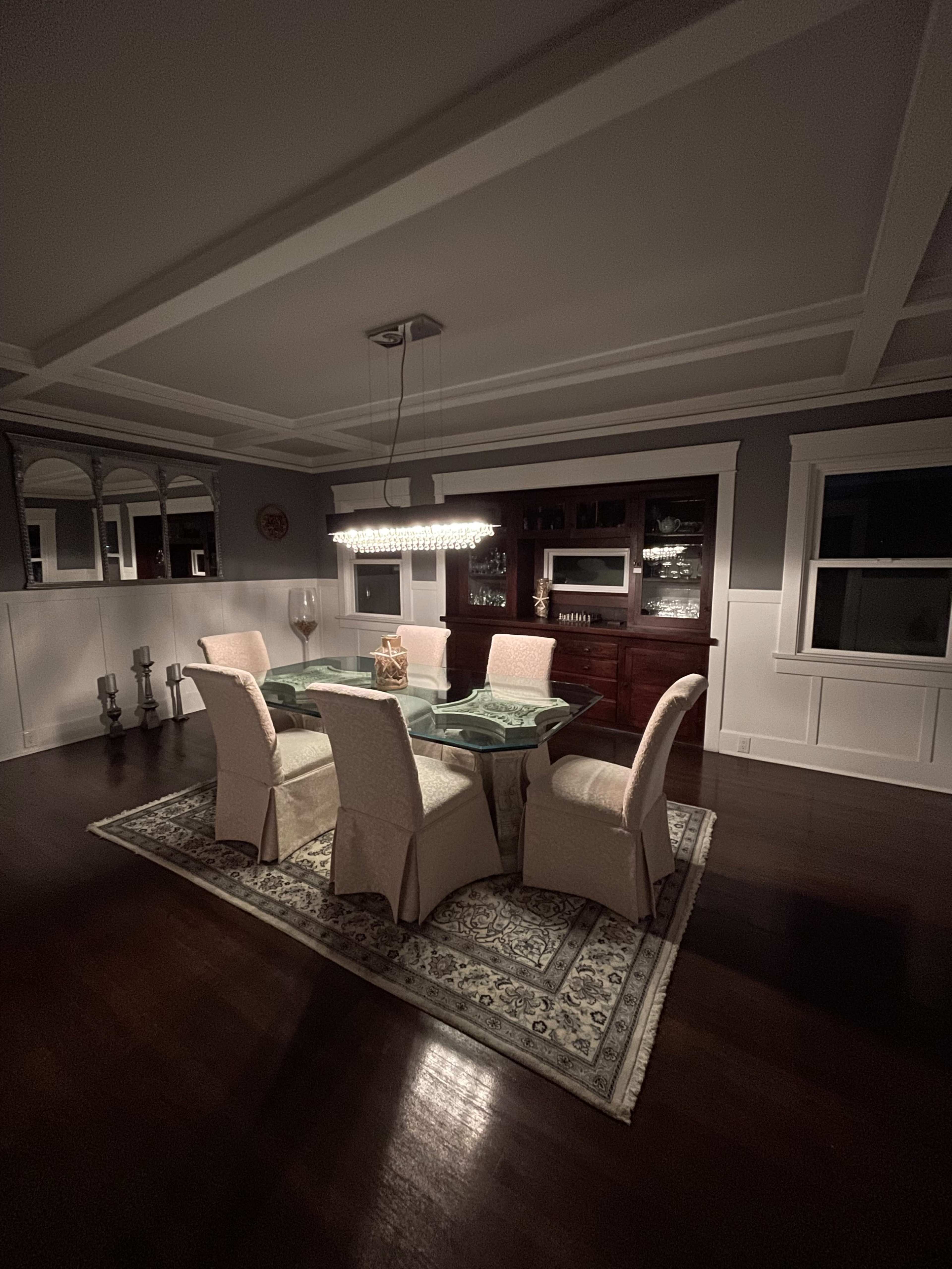 A dining area features a glass table surrounded by six upholstered chairs, illuminated by a modern ceiling light, with a sideboard and decor in the background.