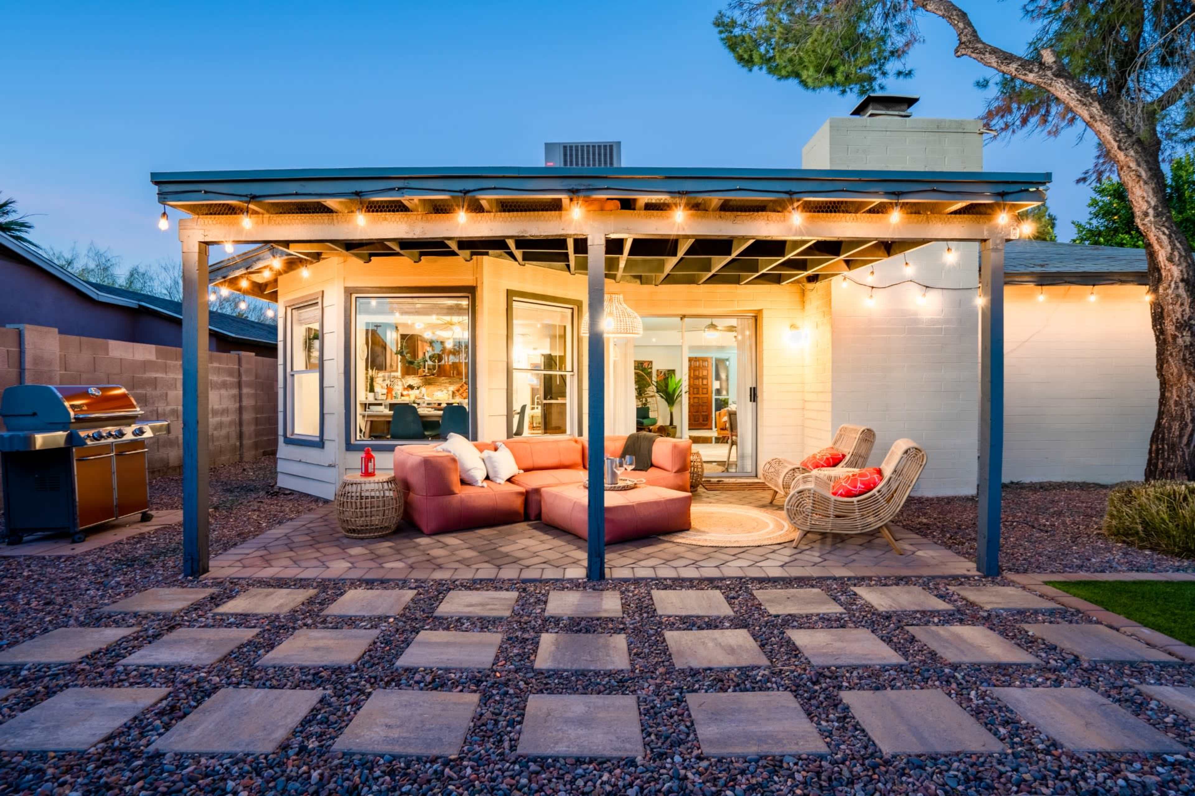 A cozy outdoor patio area with a sectional sofa, two chairs, and a grill, surrounded by landscaping and illuminated by string lights.