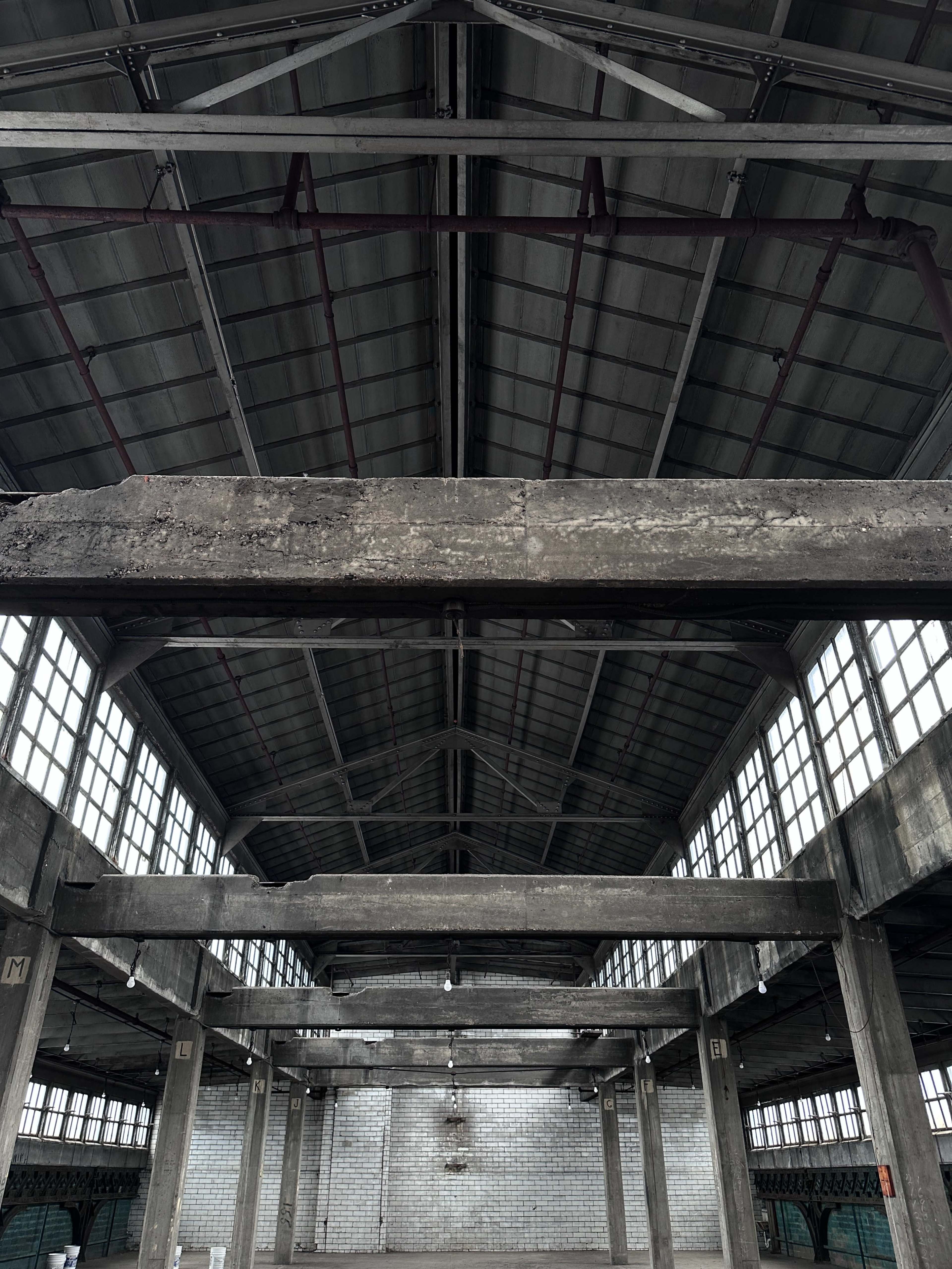 The image shows the interior of a large, industrial space with high ceilings, exposed beams, and large windows allowing natural light to enter.
