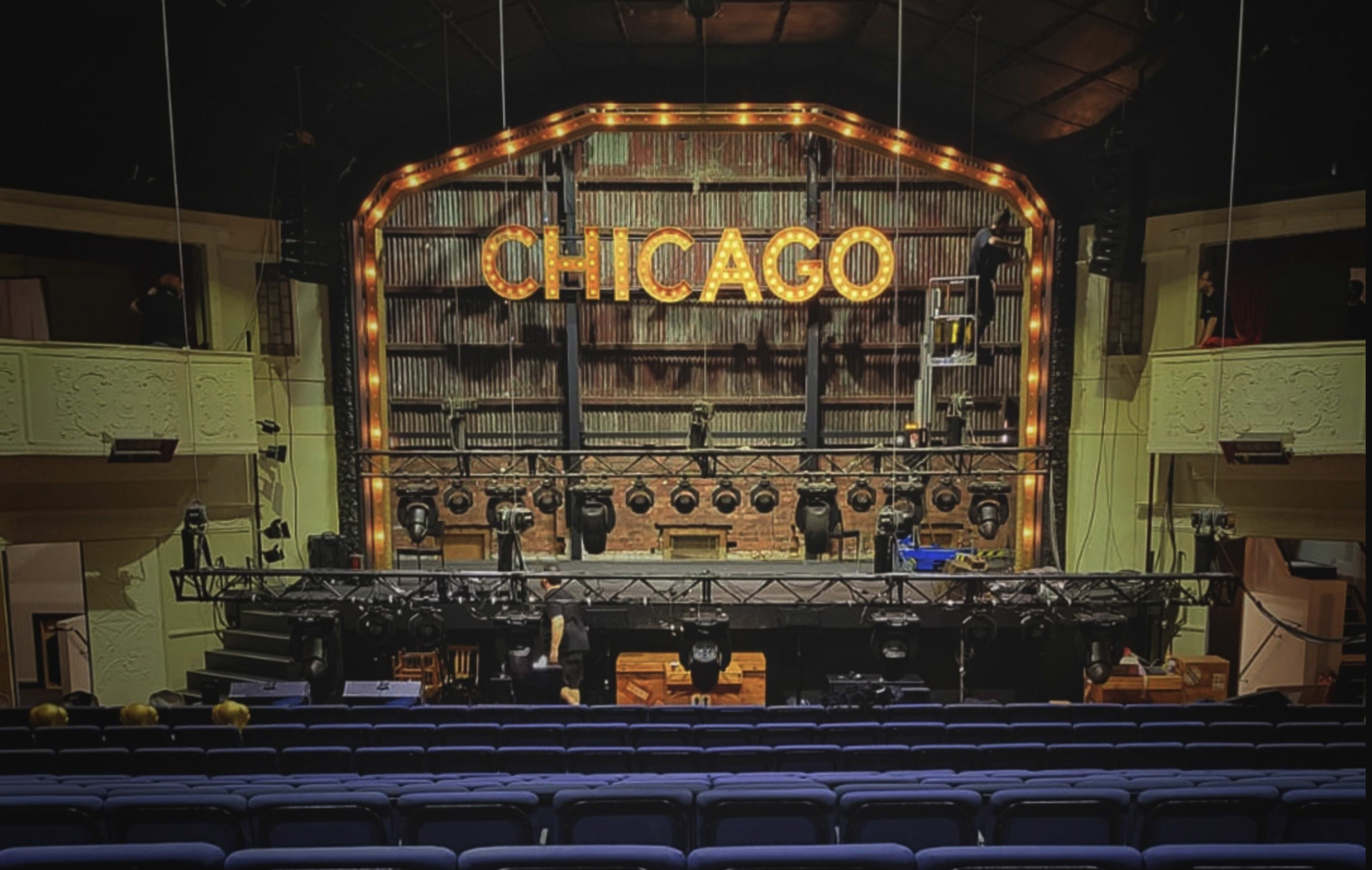 The stage of a theater is set for a performance of "Chicago," featuring illuminated letters above the stage and empty blue seats in the foreground.