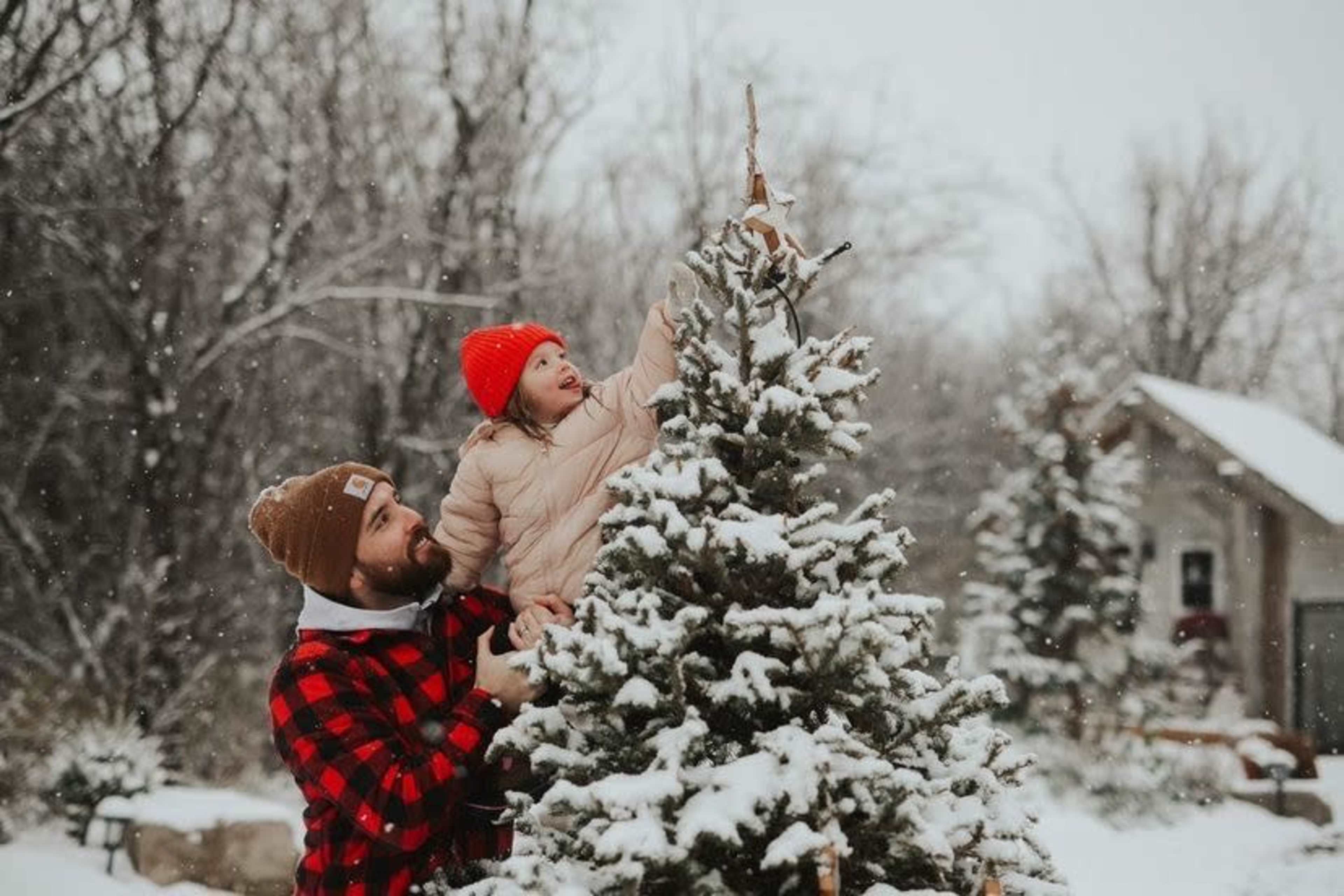 A man helps a young girl place a star on a snow-covered Christmas tree in a snowy outdoor setting.