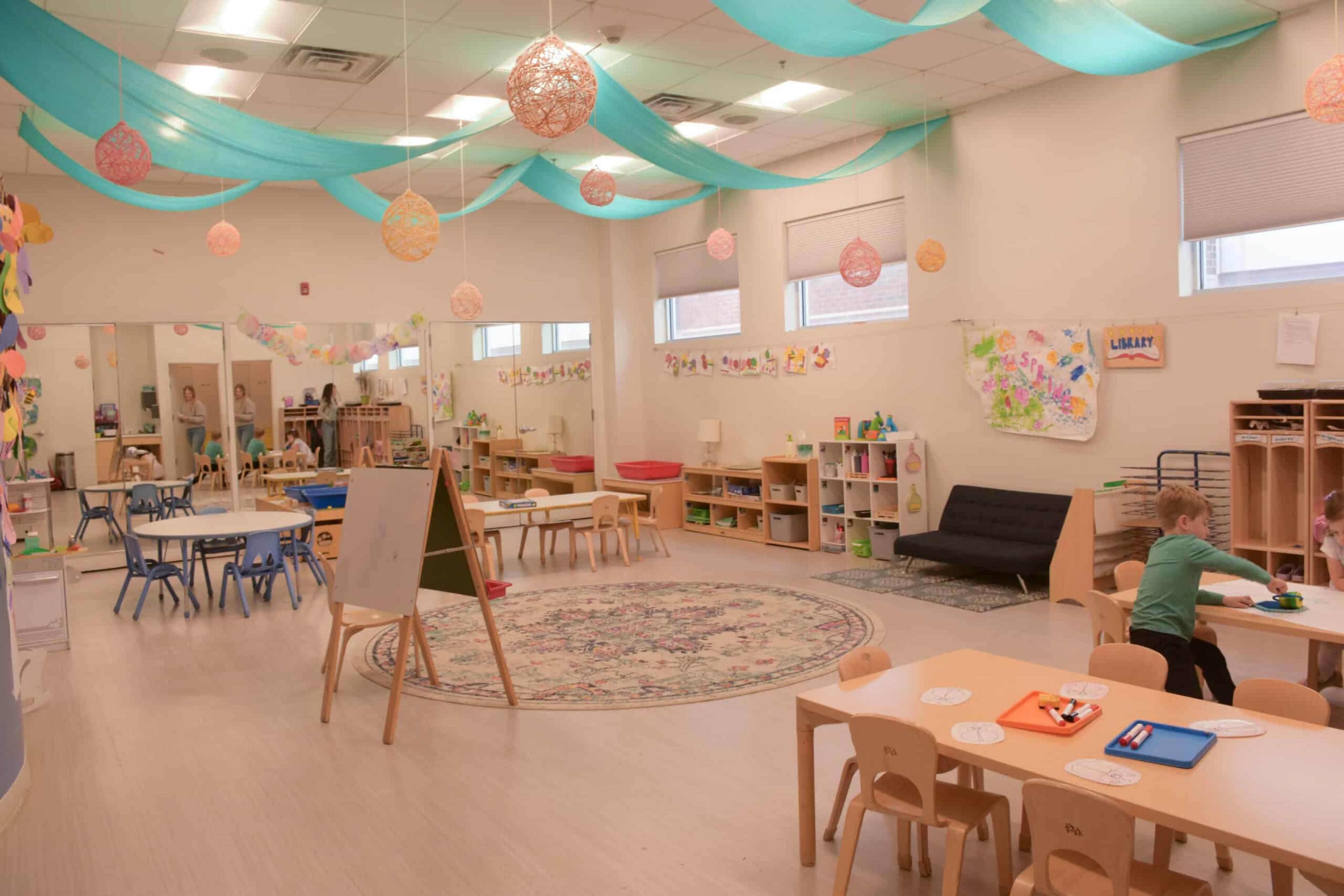 A brightly lit classroom features colorful decorations, tables for activities, and shelves lined with educational materials.