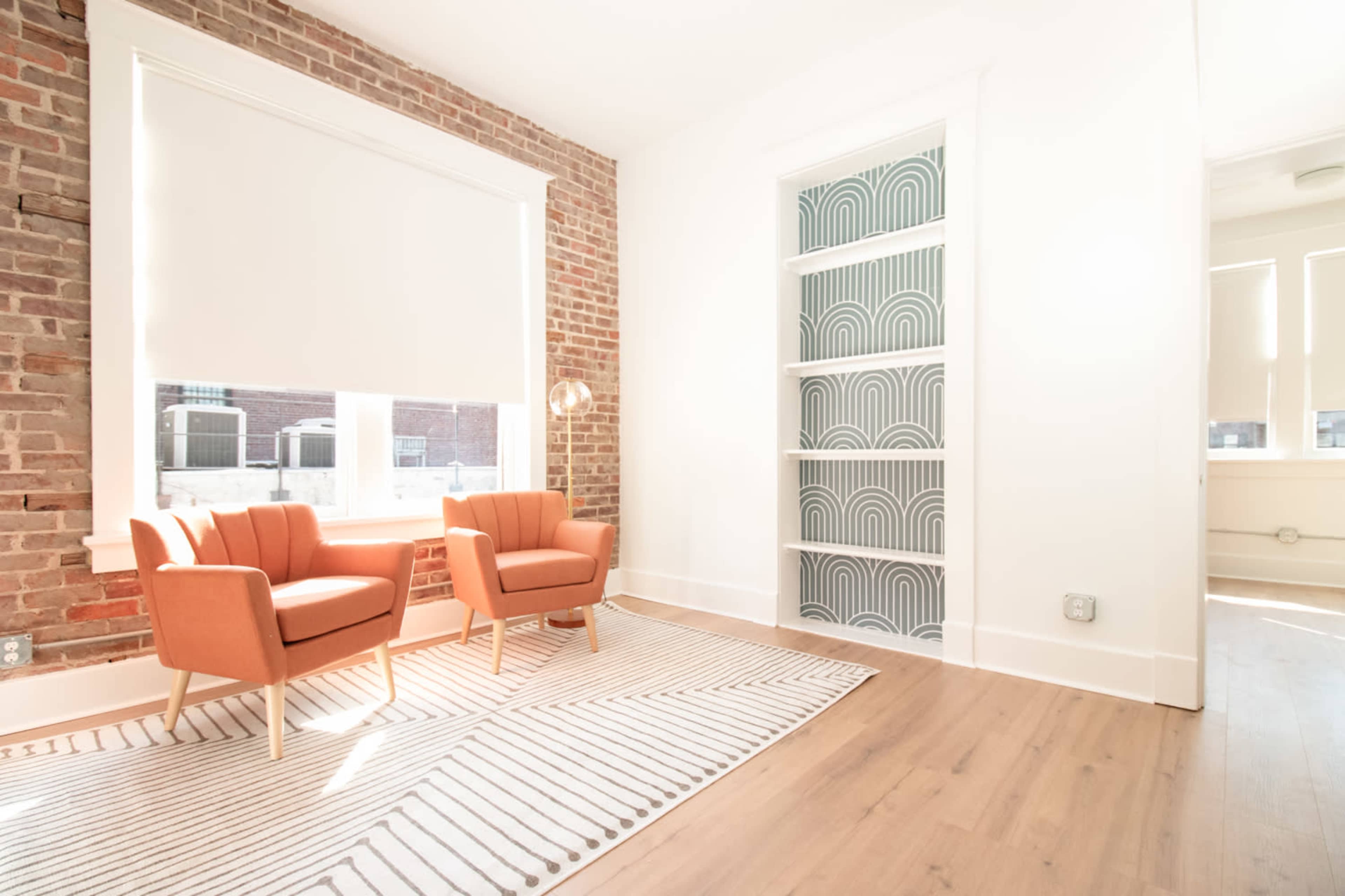 A well-lit room featuring two orange chairs, a patterned rug, and a bookshelf with an arch design against a brick wall.