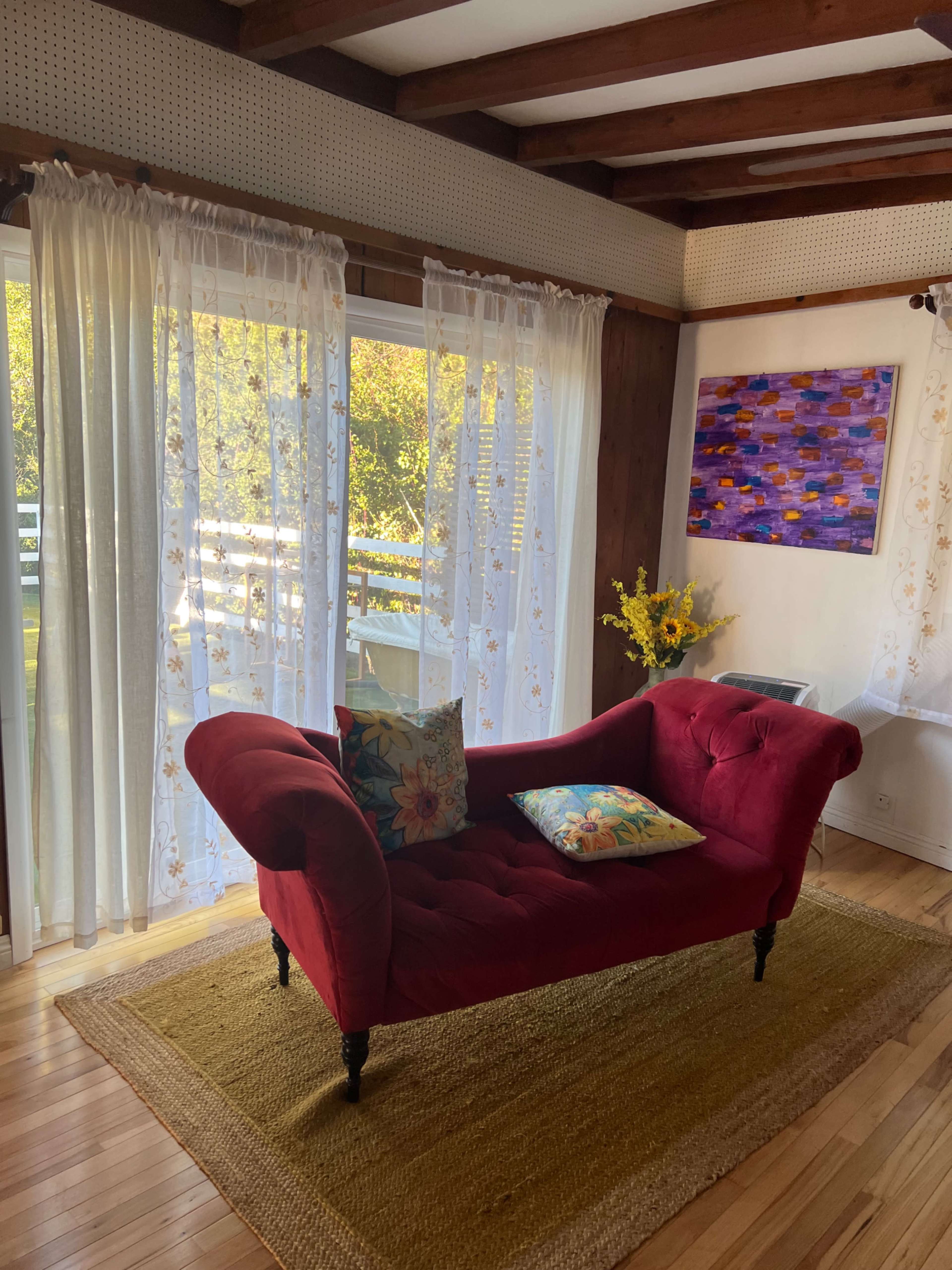 A red chaise lounge with decorative pillows is positioned near large windows draped with sheer curtains, overlooking a serene outdoor view, with a piece of artwork hanging on the wall.