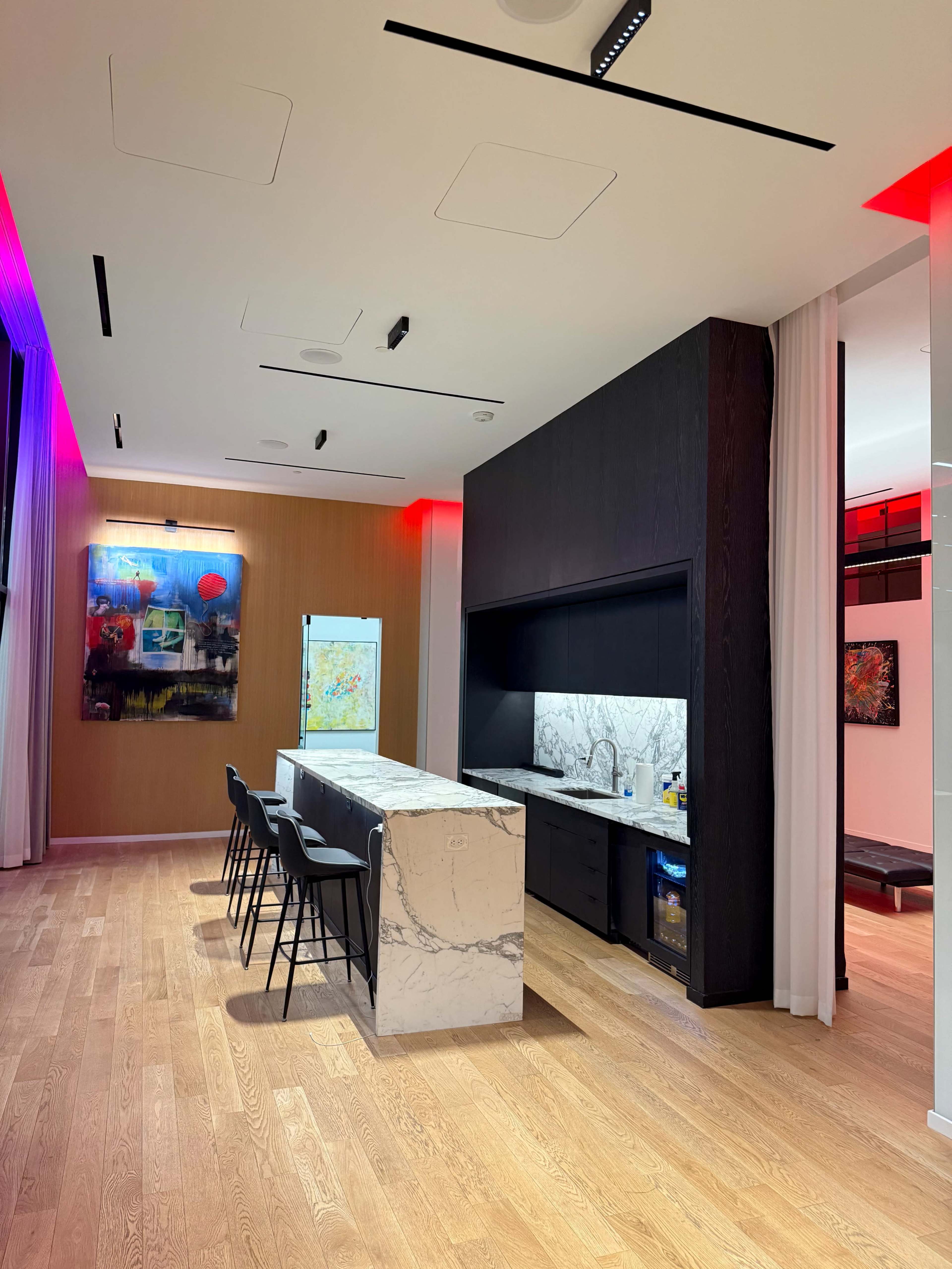 The image shows a modern kitchen area with a marble island, black cabinetry, wooden flooring, and colorful artwork on the walls under ambient lighting.