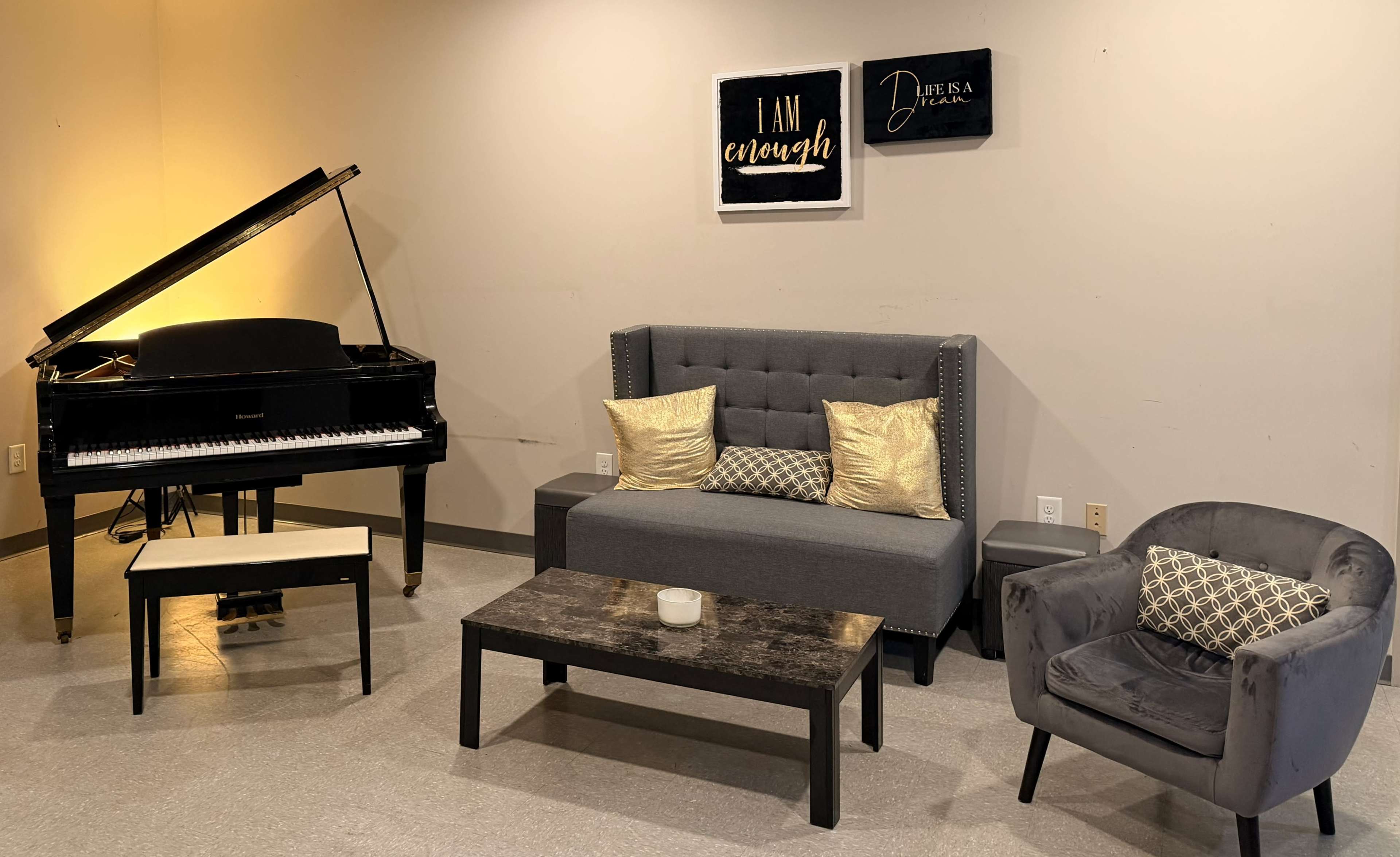 The image shows a room with a black grand piano on the left, a gray couch centered in the middle, a black coffee table in front of it, and a gray accent chair to the right, all set against a plain wall with framed decorative quotes above the couch.