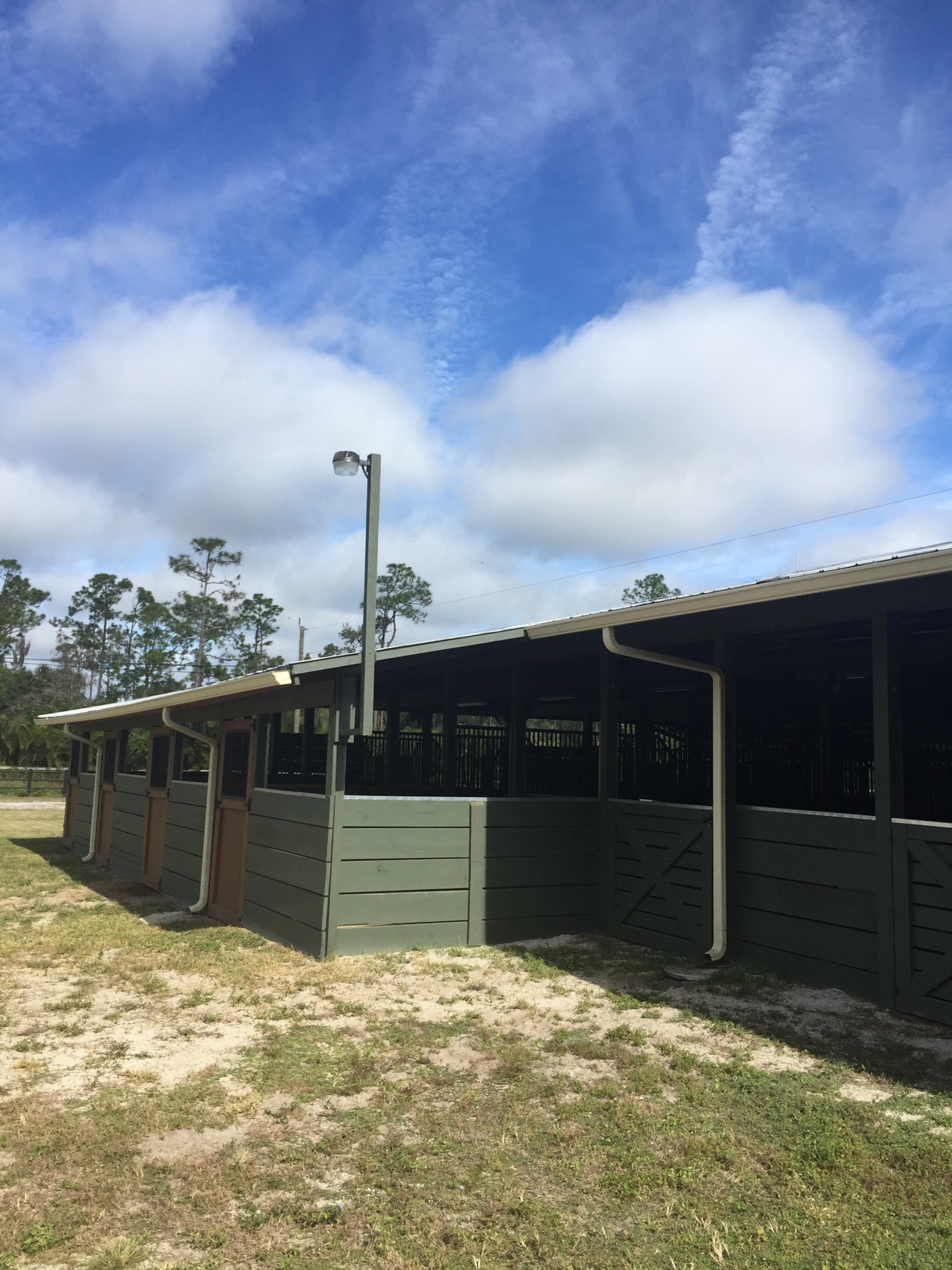 A row of horse stalls is lined along a green wooden structure under a partly cloudy sky.