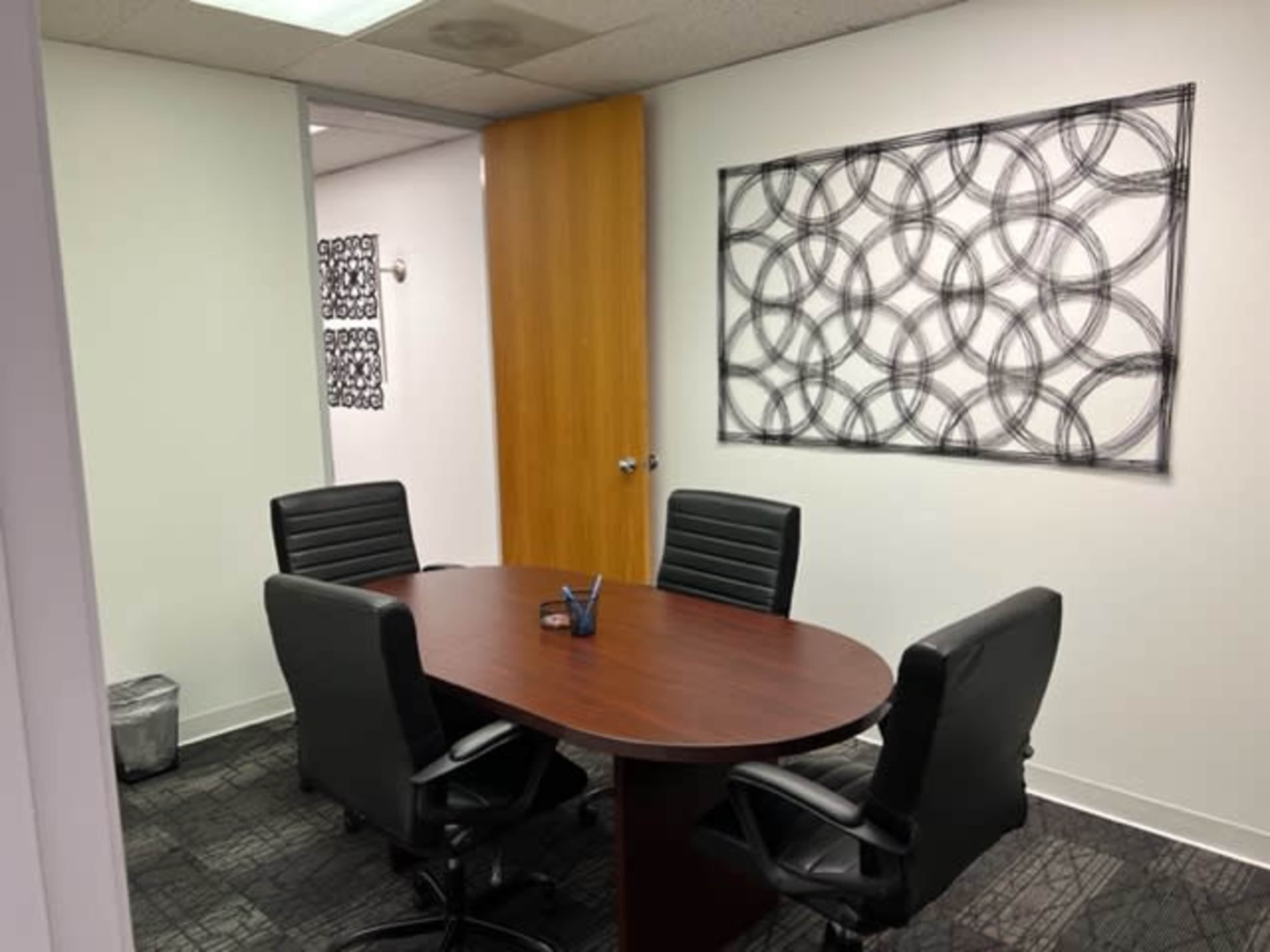 A conference room is furnished with a round wooden table and four black leather chairs, featuring a framed artwork on the wall.