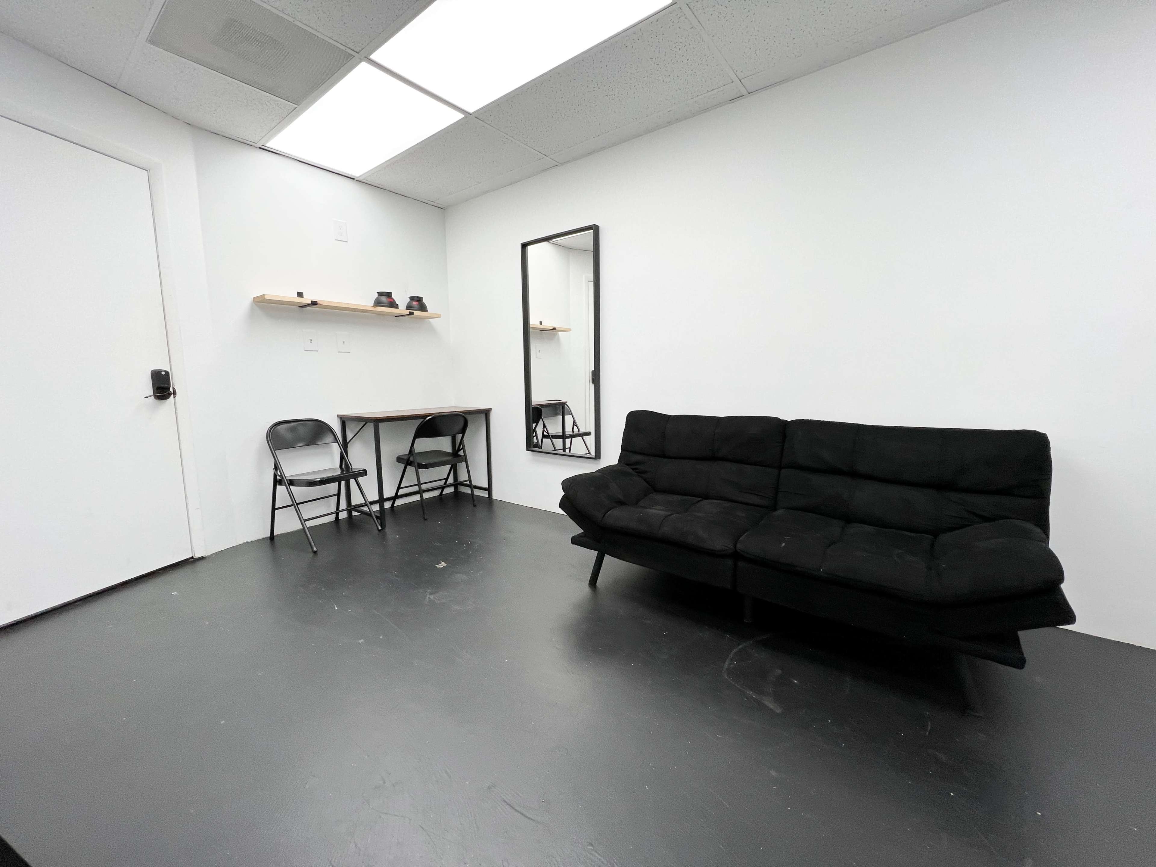 A minimalistic room with a black sofa, a small table with two chairs, and a mirror on a white wall.