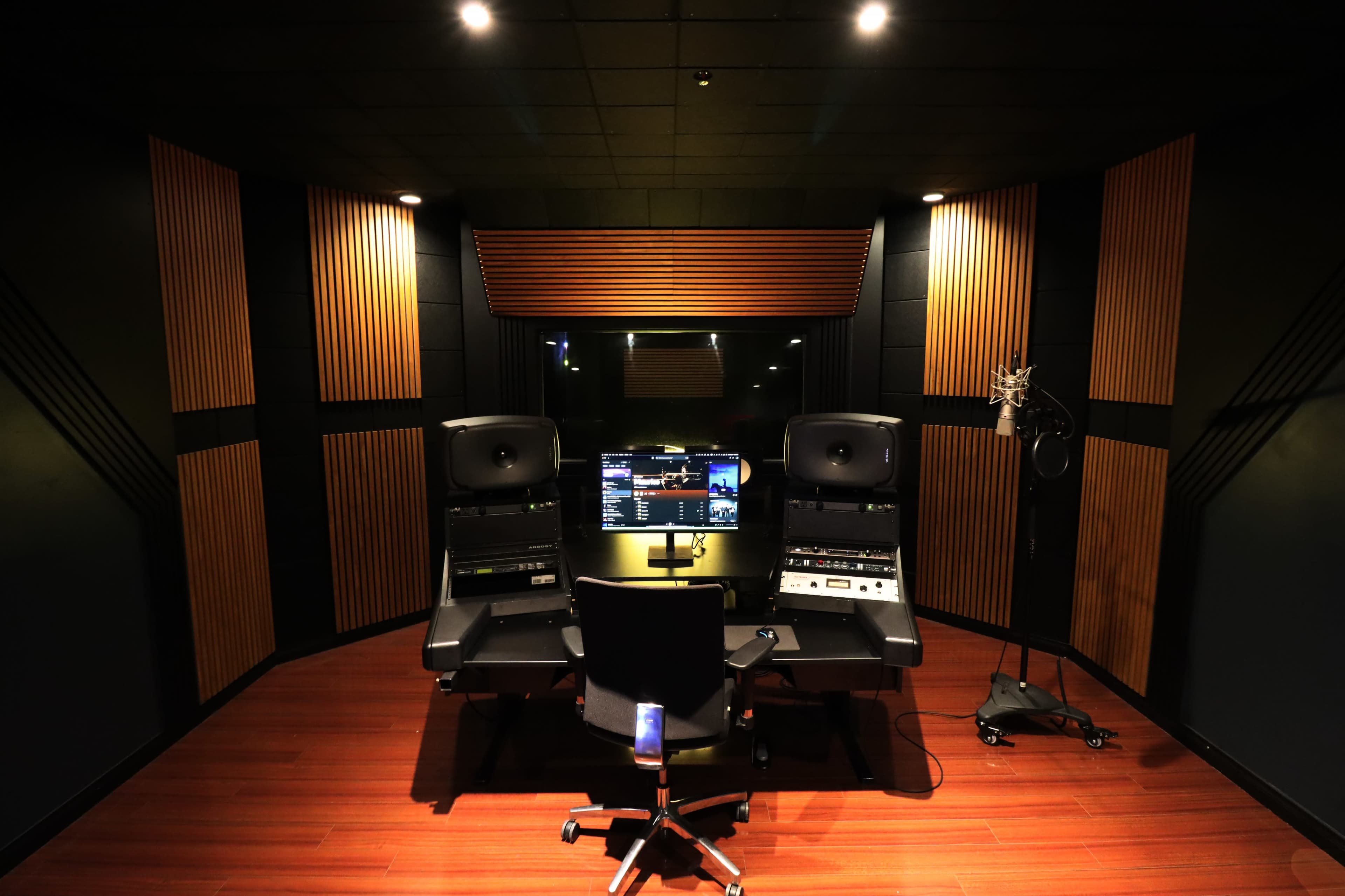 The image shows a recording studio with a central workspace featuring two large speakers, a computer monitor, and a microphone stand in a soundproof room with wooden paneling.